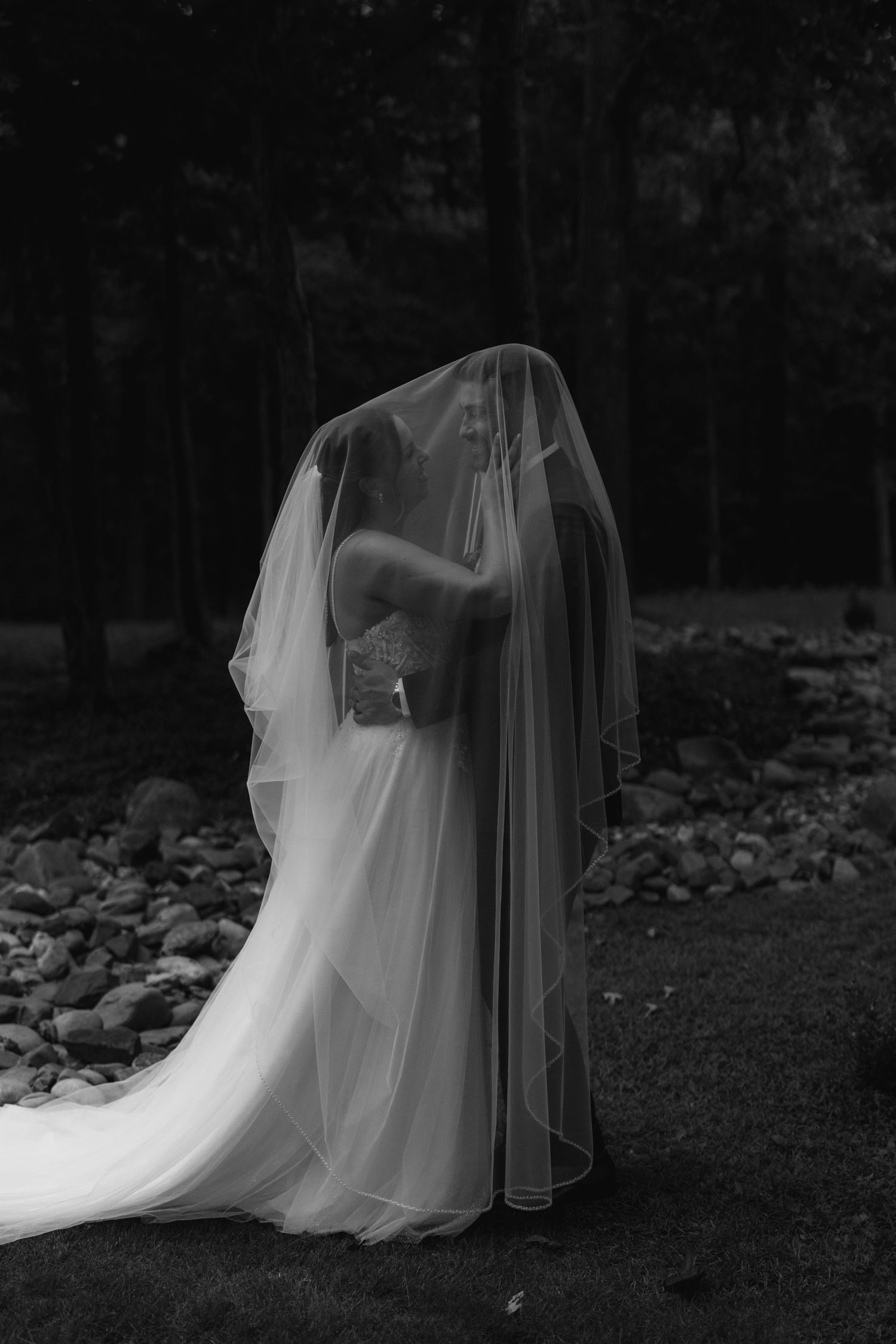 Black and white bridal portrait of a bride draped in a flowing veil outdoors among trees and rocks.