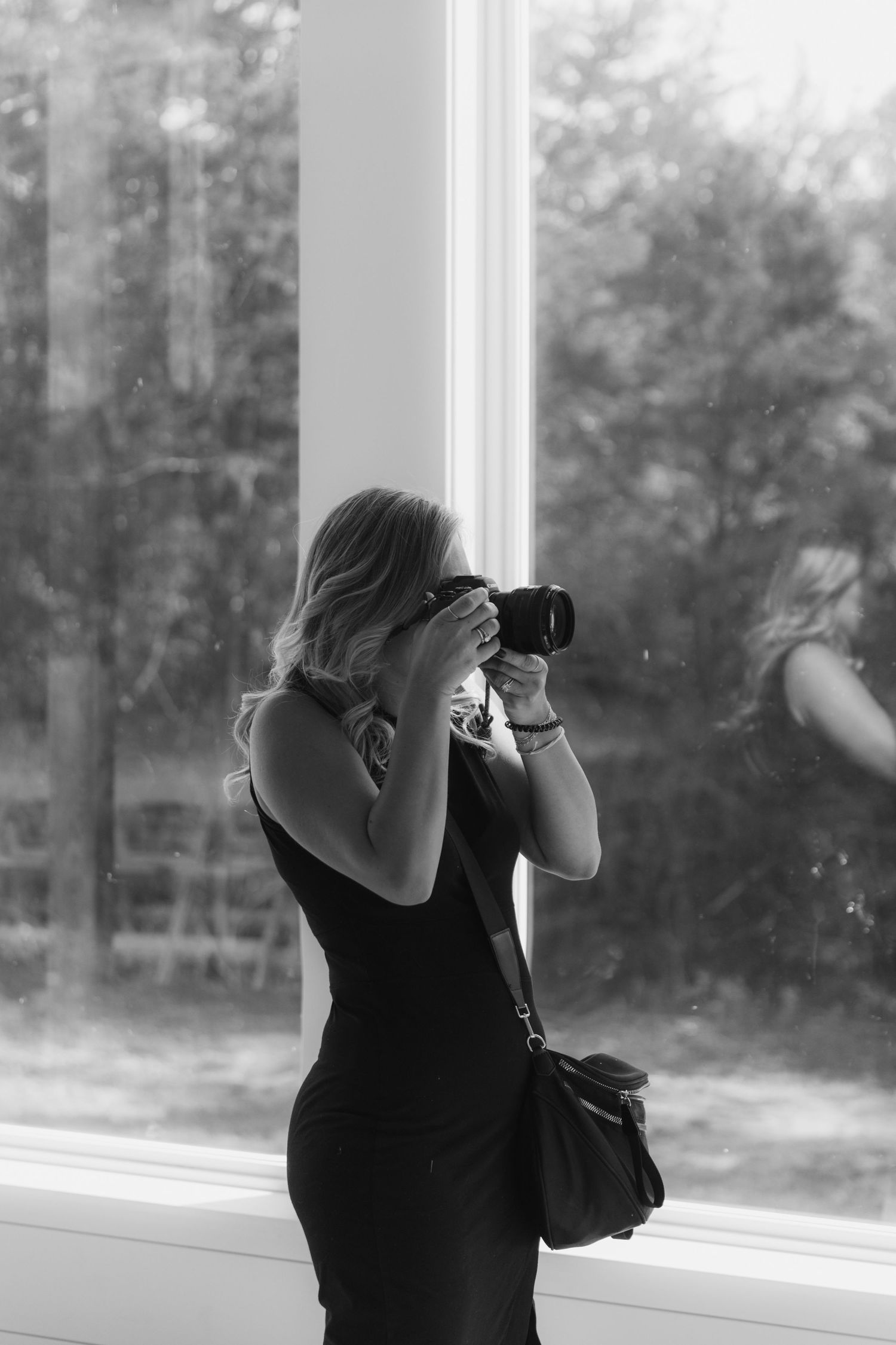 A photographer in a black top takes photos near a large window with trees visible outside.