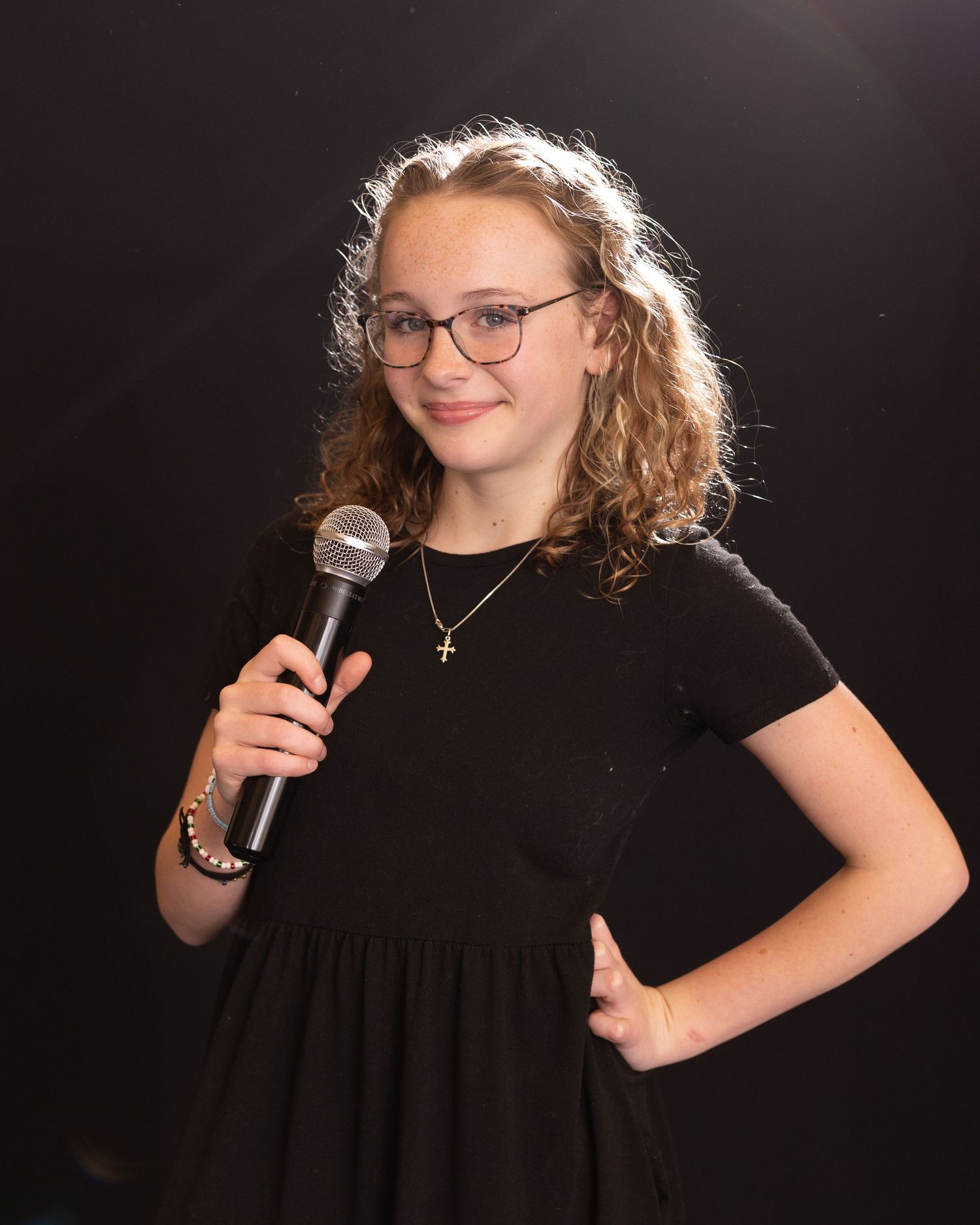 A young performer in a black dress and glasses holds a microphone while posing against a dark background in multiple poses.