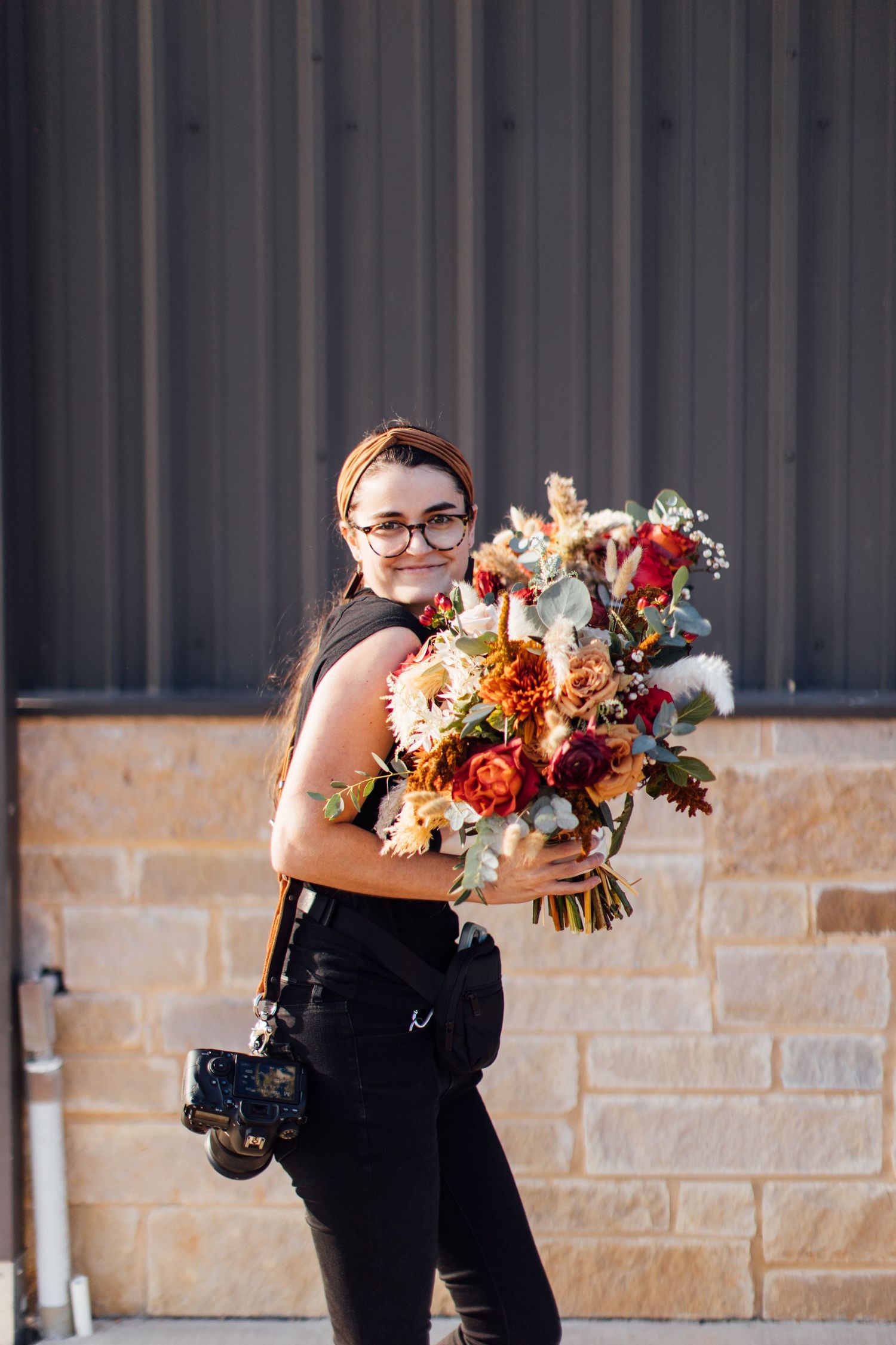 A photographer in black clothing carries a vibrant bouquet of colorful flowers against a stone and metal wall backdrop.