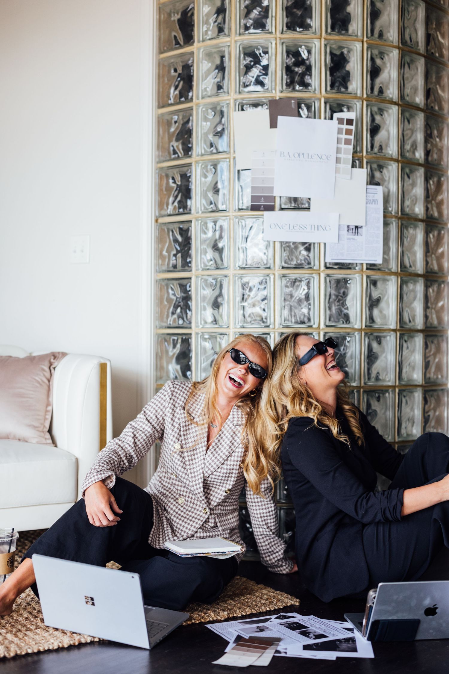 Two stylish women in sunglasses laughing while sitting on floor surrounded by design materials and laptops.