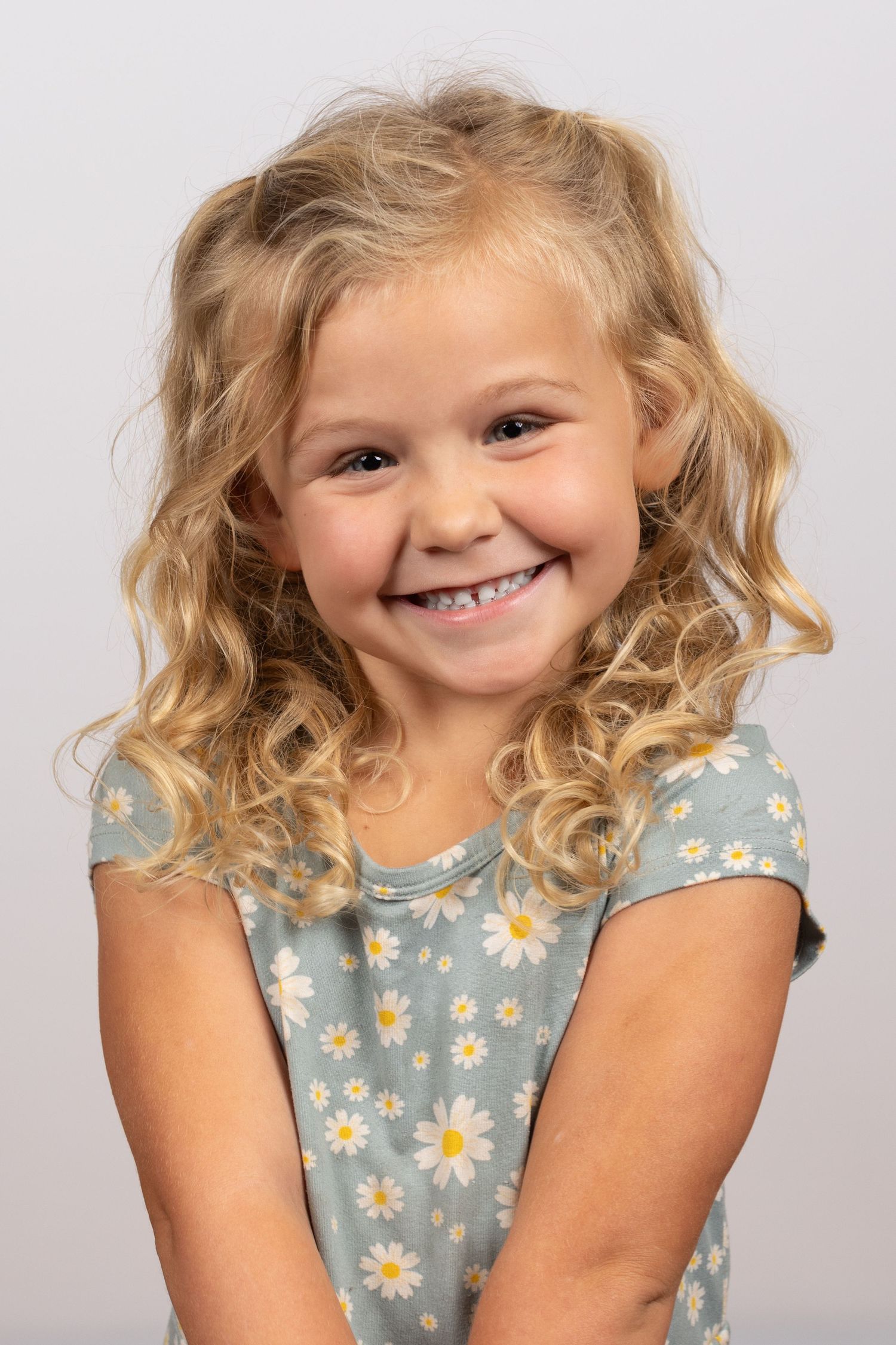 A child with curly blonde hair wearing a blue floral dress smiles brightly against a light background.