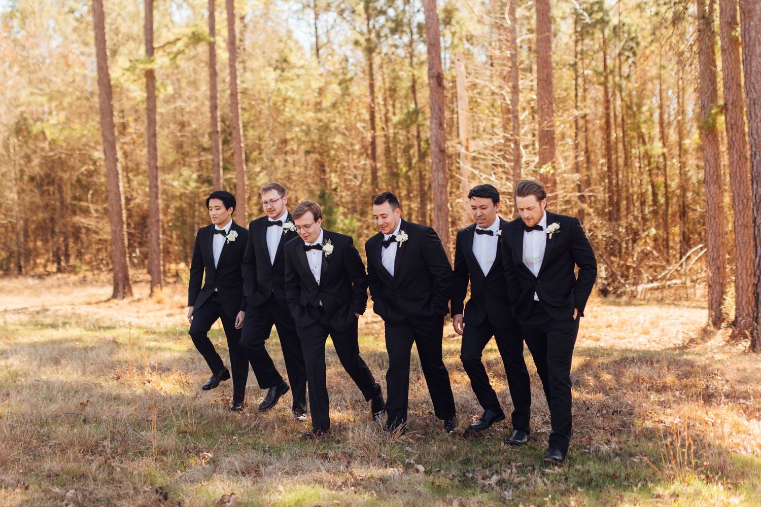 Groomsmen in black tuxedos pose together in wooded outdoor setting.