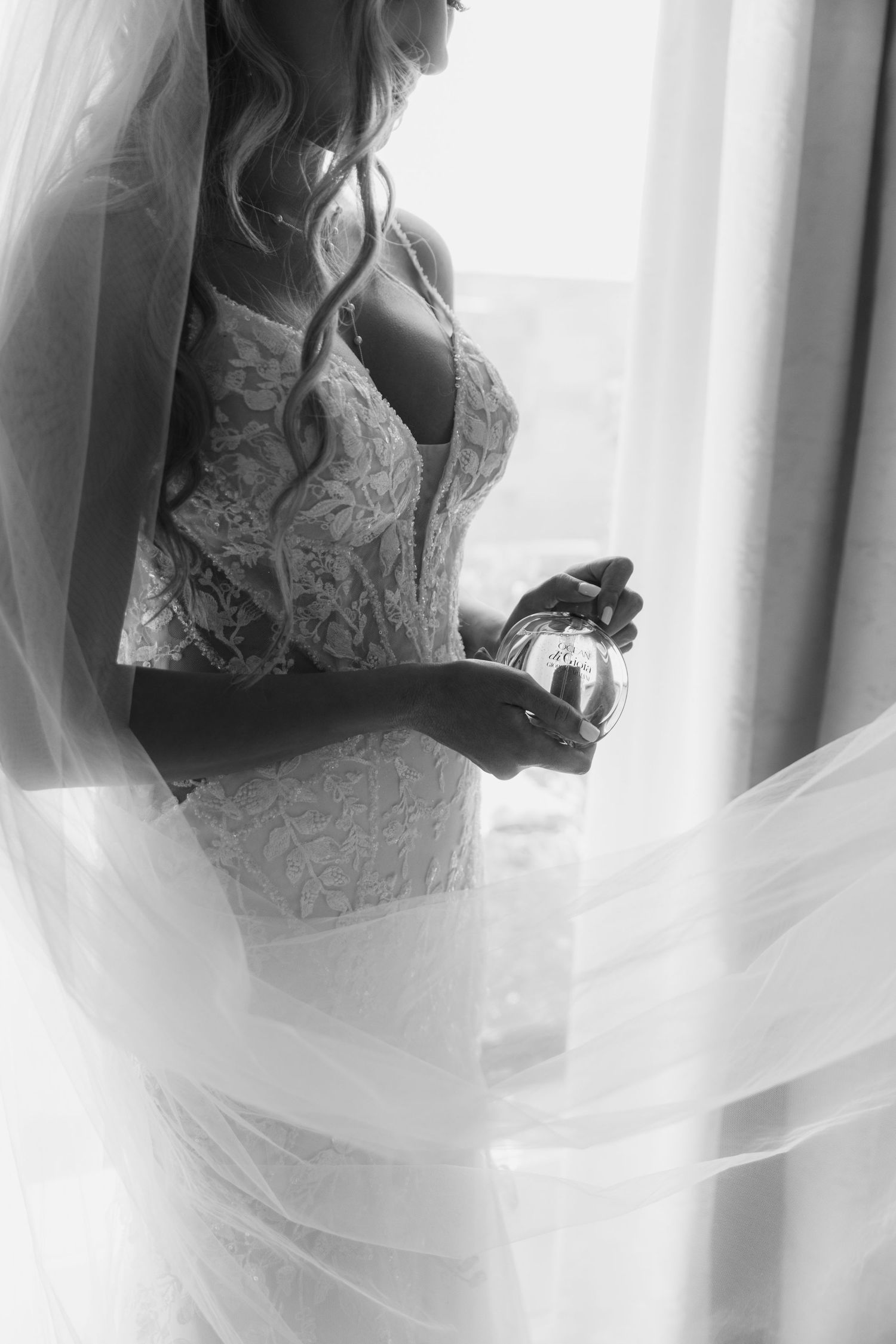 Black and white portrait of a bride in lace dress holding a perfume bottle next to a window.