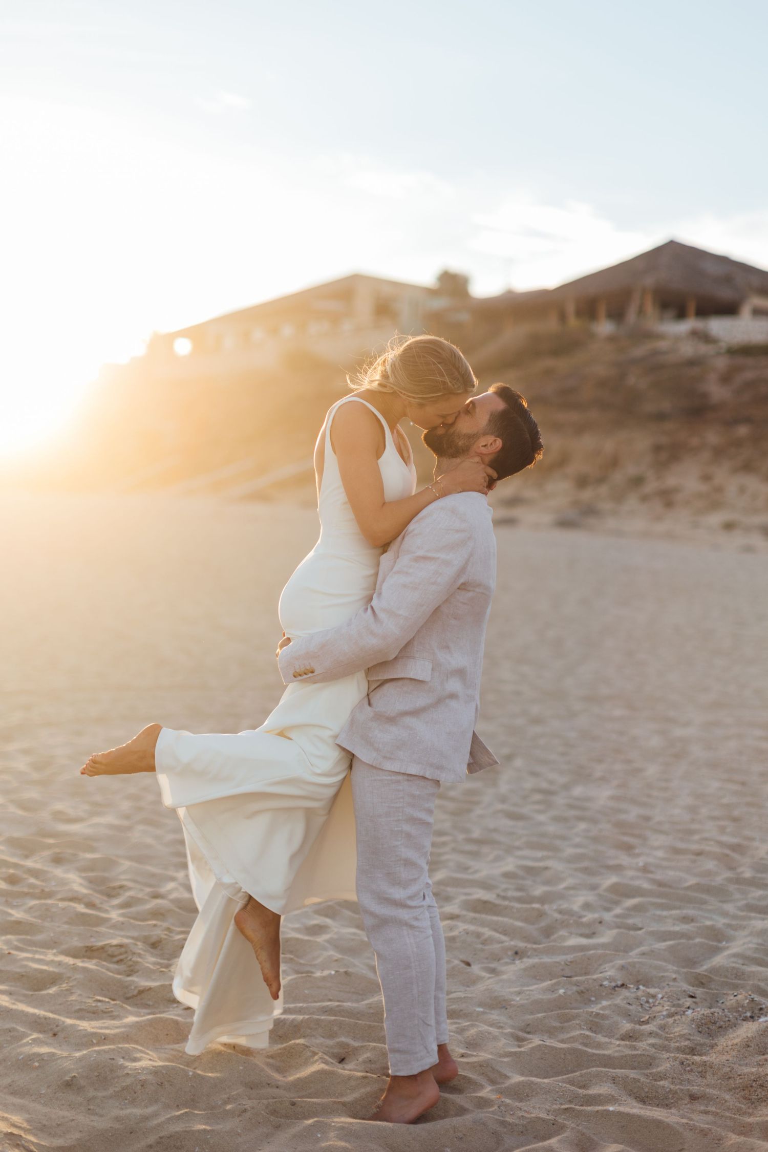 Couple in white outfits embracing on sandy beach during golden hour sunset.