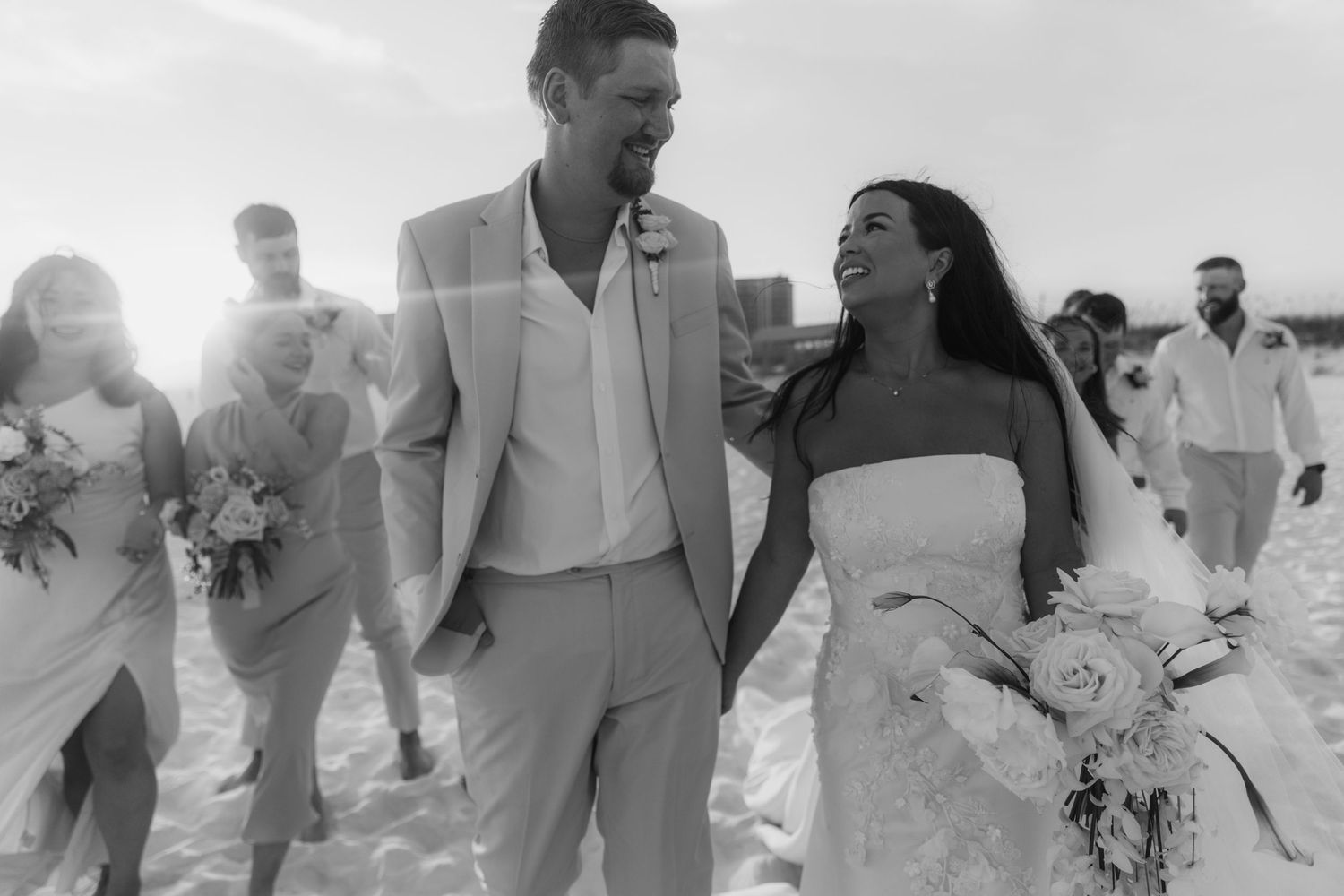 Black and white beach wedding photo of smiling couple holding hands walking with bridal party in sunny outdoor ceremony.