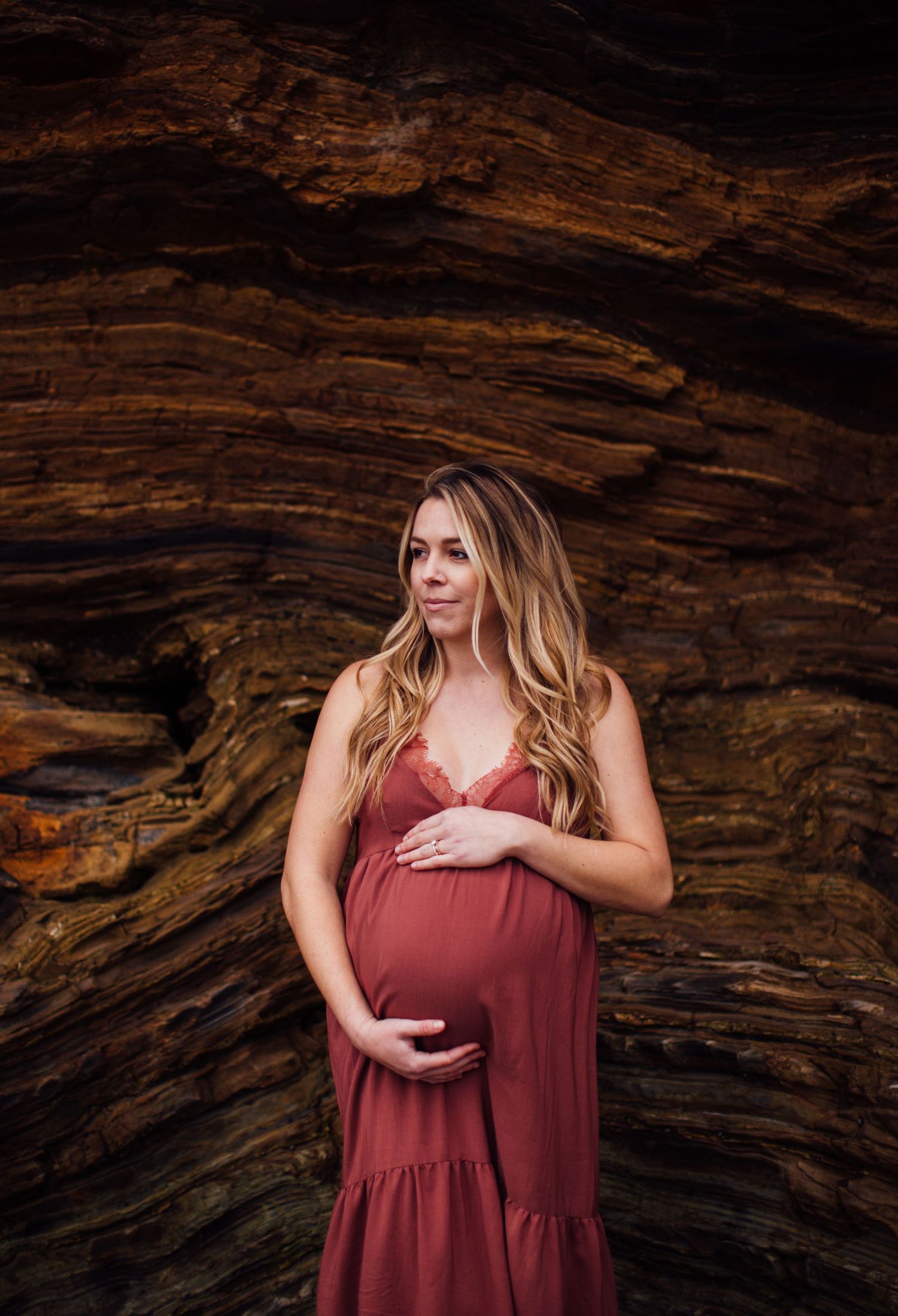 Pregnant woman in a rust-colored maternity dress cradling her belly against layered rock formations.