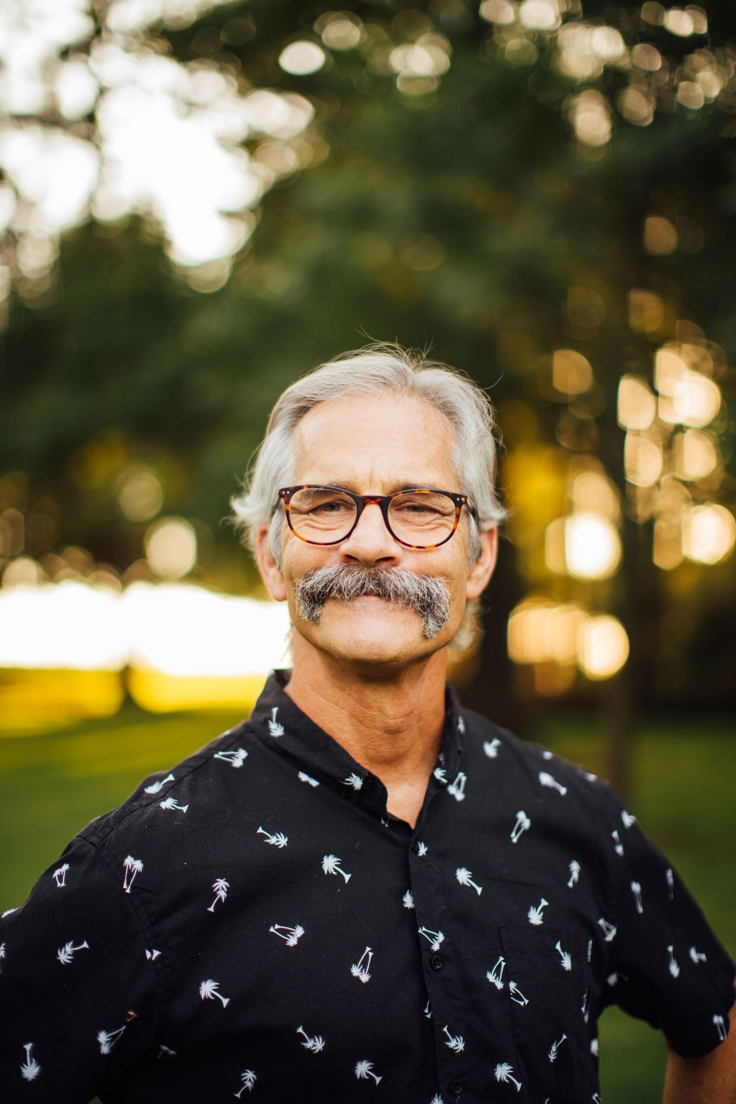 Smiling person with gray hair and mustache wearing glasses and a patterned short-sleeve button-up shirt outdoors at sunset.