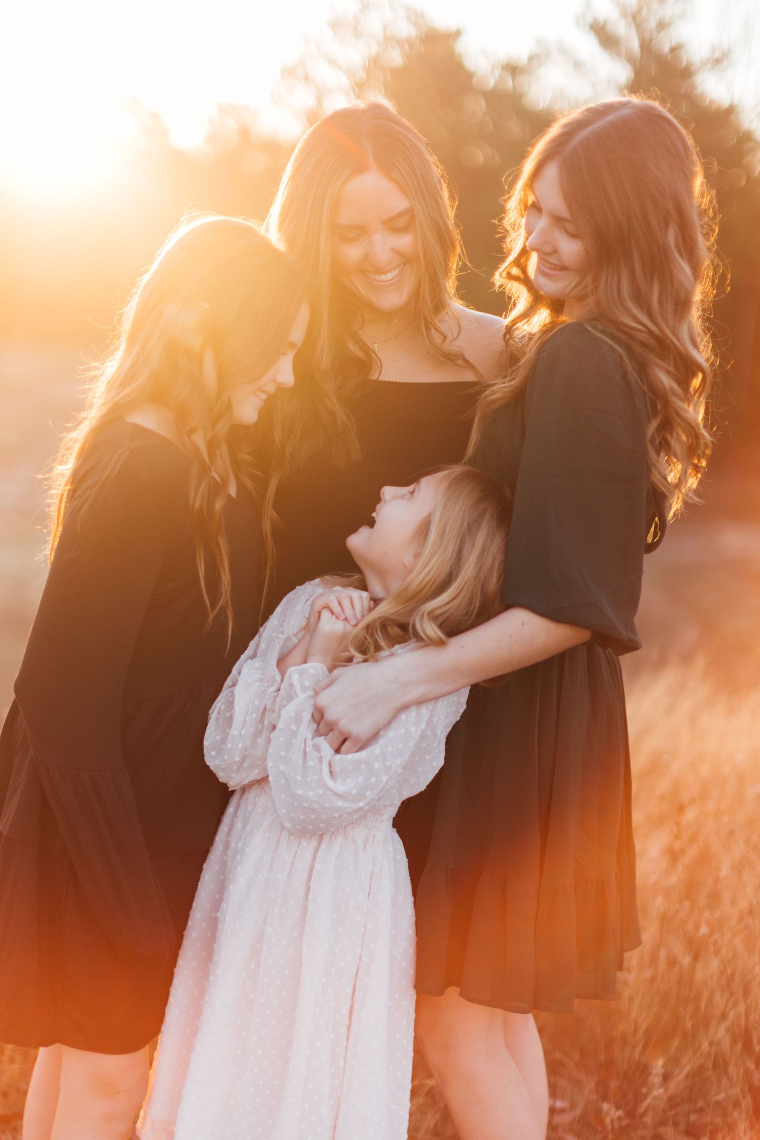 Three women in black and white dresses share a tender moment in warm golden sunlight at sunset.