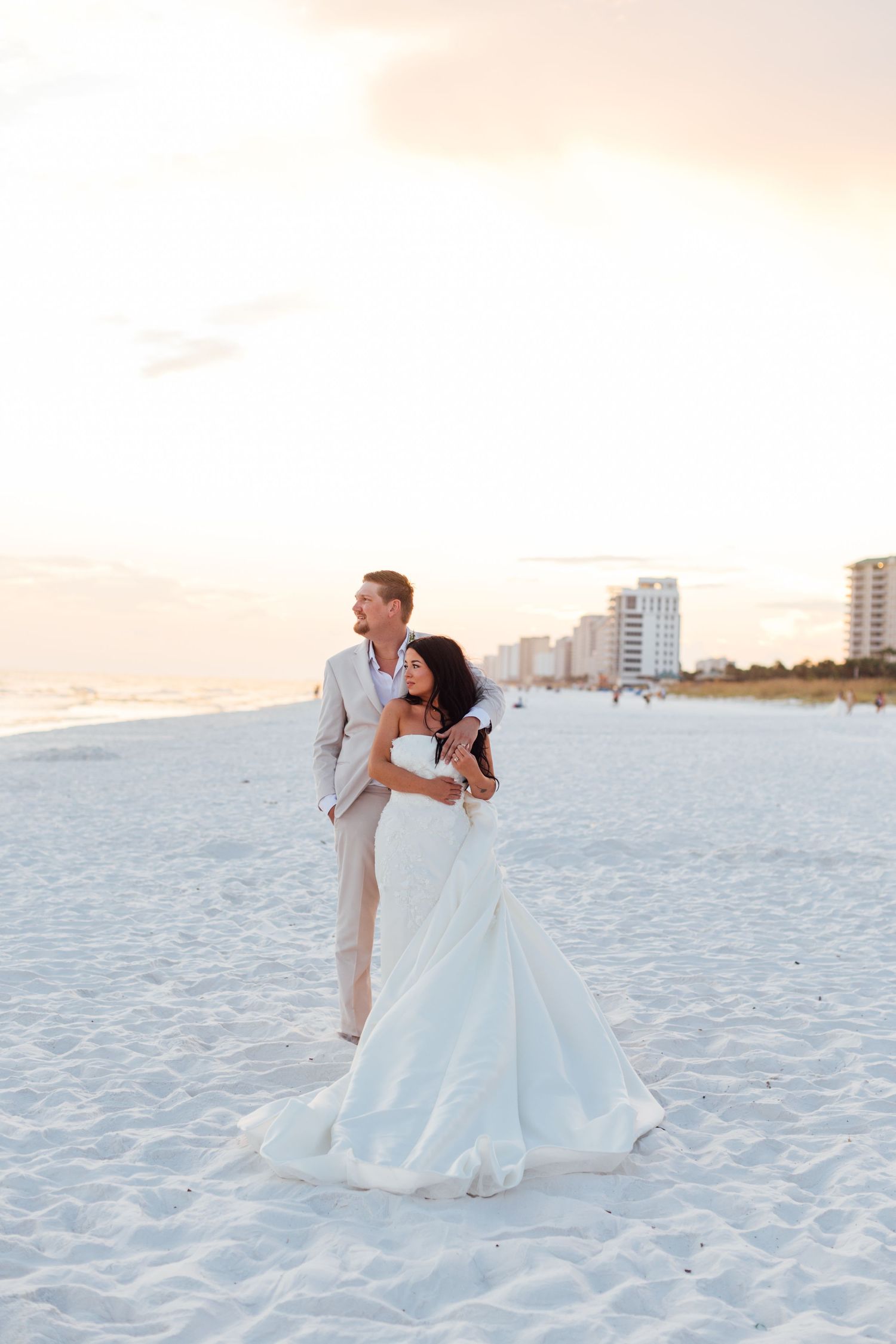 Wedding photography and groom embrace on a white sand beach at sunset, with the bride in a flowing white gown and groom in a tan suit.