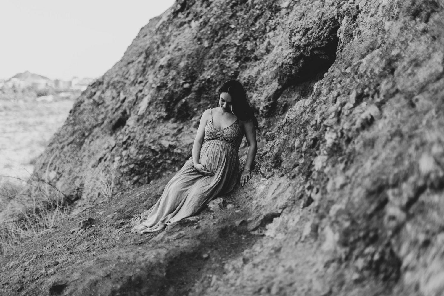 Black and white photo of a pregnant woman in a flowing dress sitting against rocky cliffs outdoors.