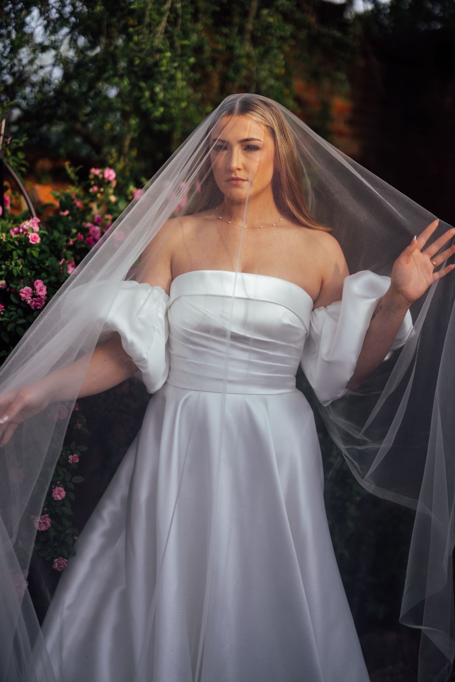Bride in strapless white satin gown with puff sleeves and flowing veil, posed among pink roses outdoors.