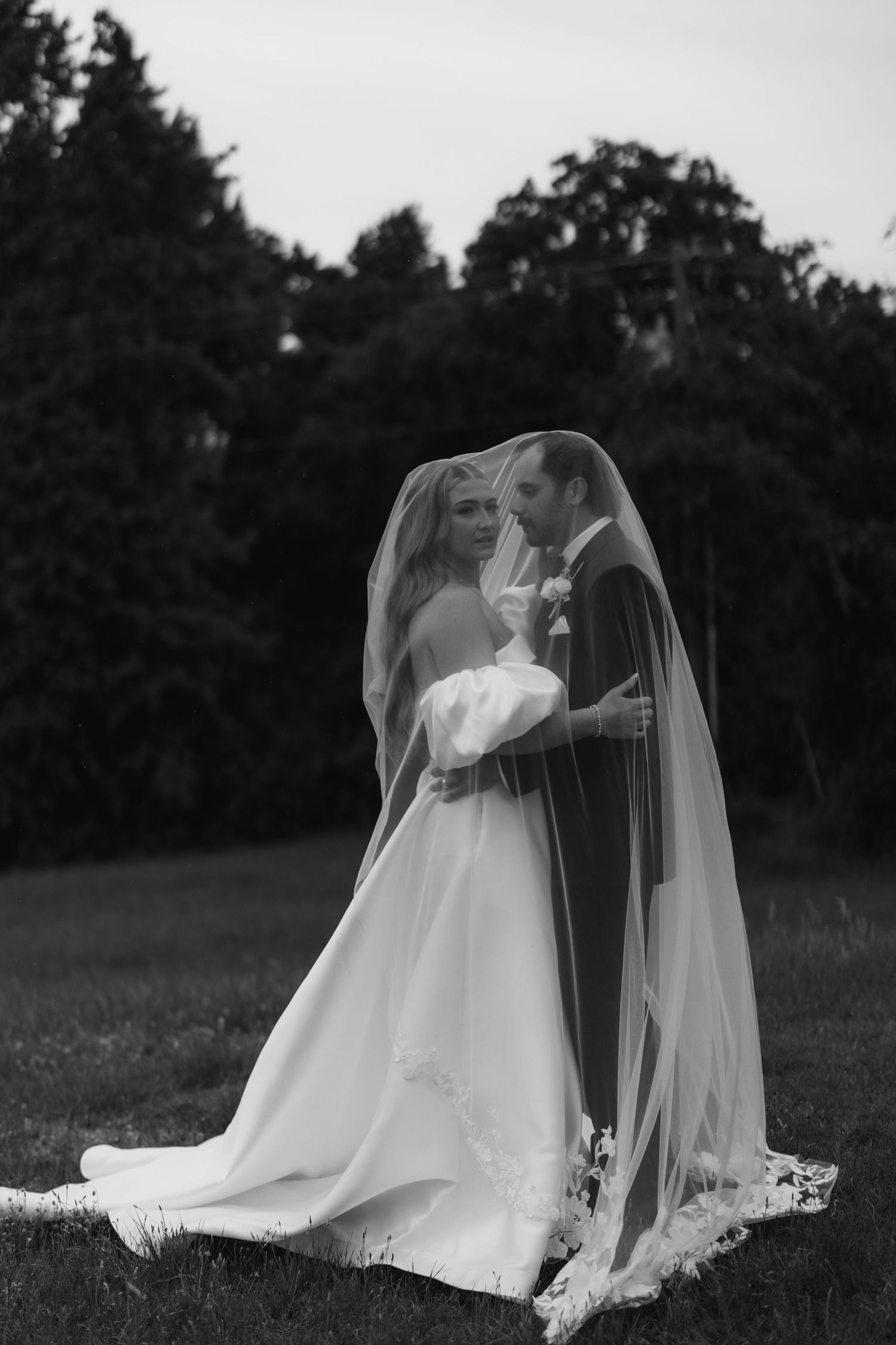 Black and white wedding photo of bride and groom sharing a romantic moment under a flowing veil outdoors.