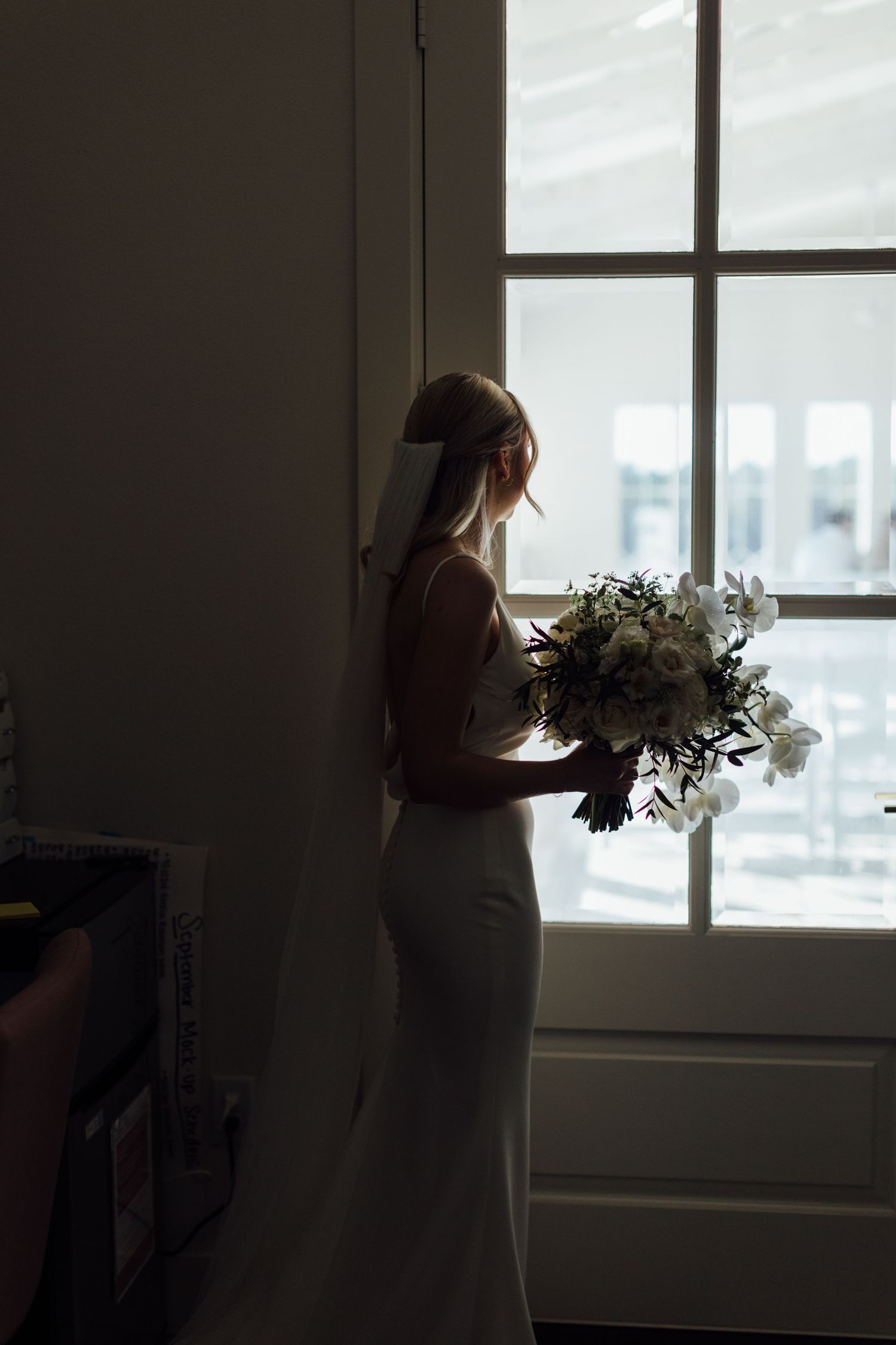 Silhouetted figure with bouquet stands in front of bright windows creating dramatic wedding portrait.