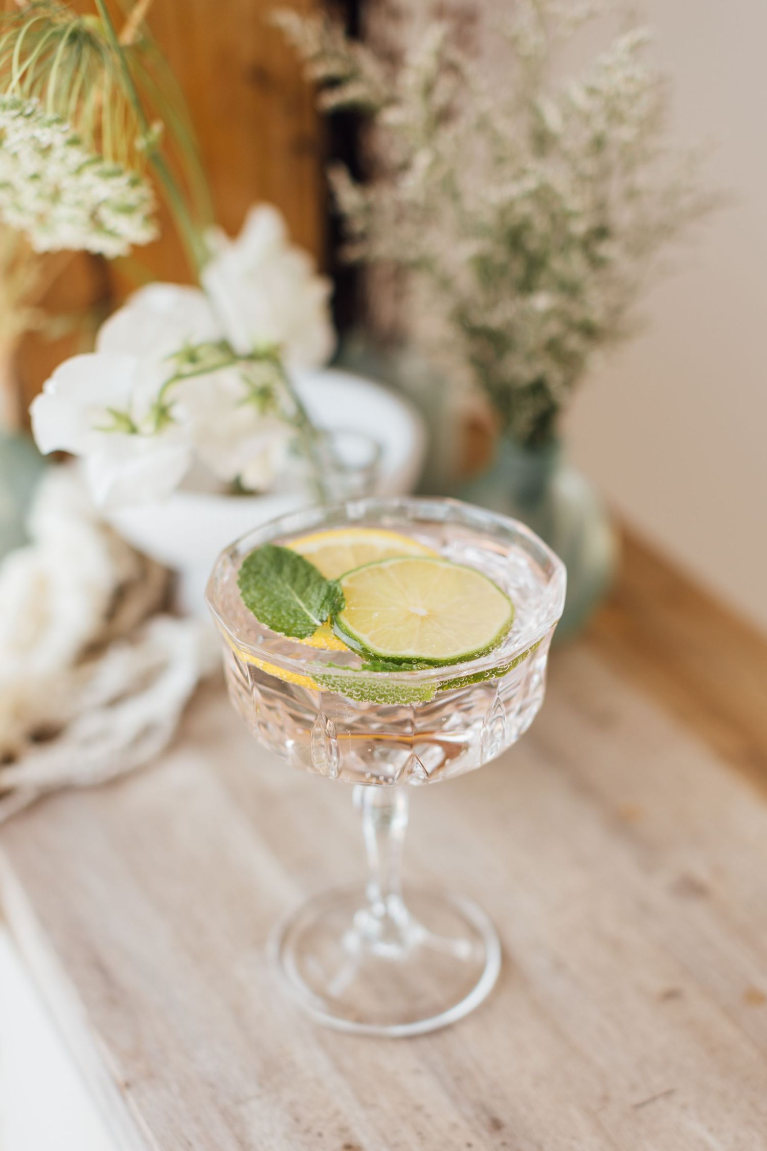 Elegant coupe cocktail glass with yellow citrus drink garnished with fresh cucumber and mint on wooden table.