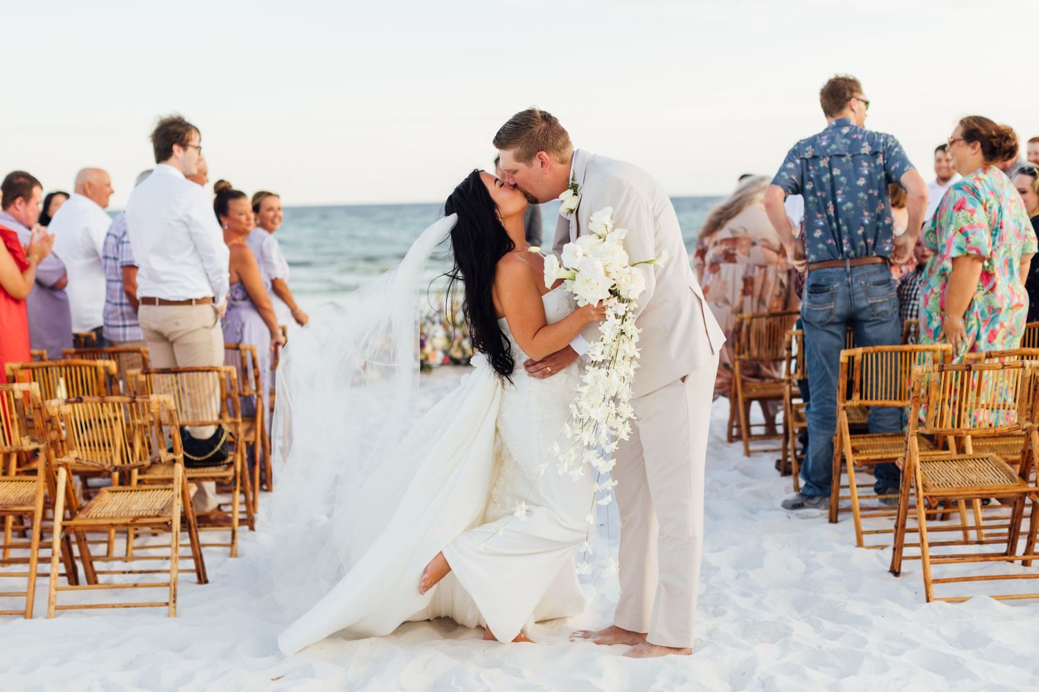 Wedding photography and groom share a kiss on a sandy beach wedding ceremony with bamboo chairs and ocean backdrop.