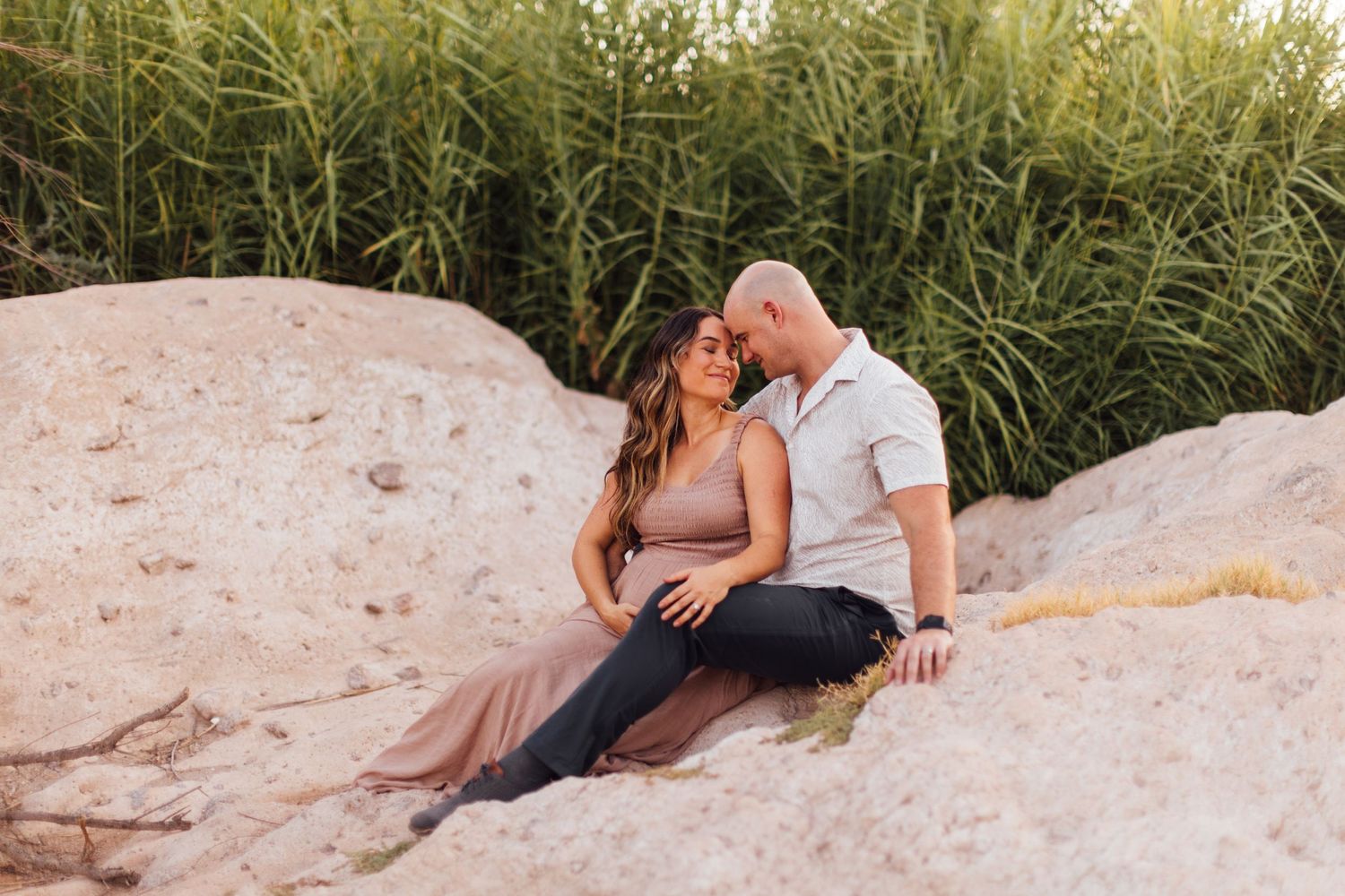 Couple sitting on large rocks sharing a romantic moment with lush green reeds in the background during golden hour.