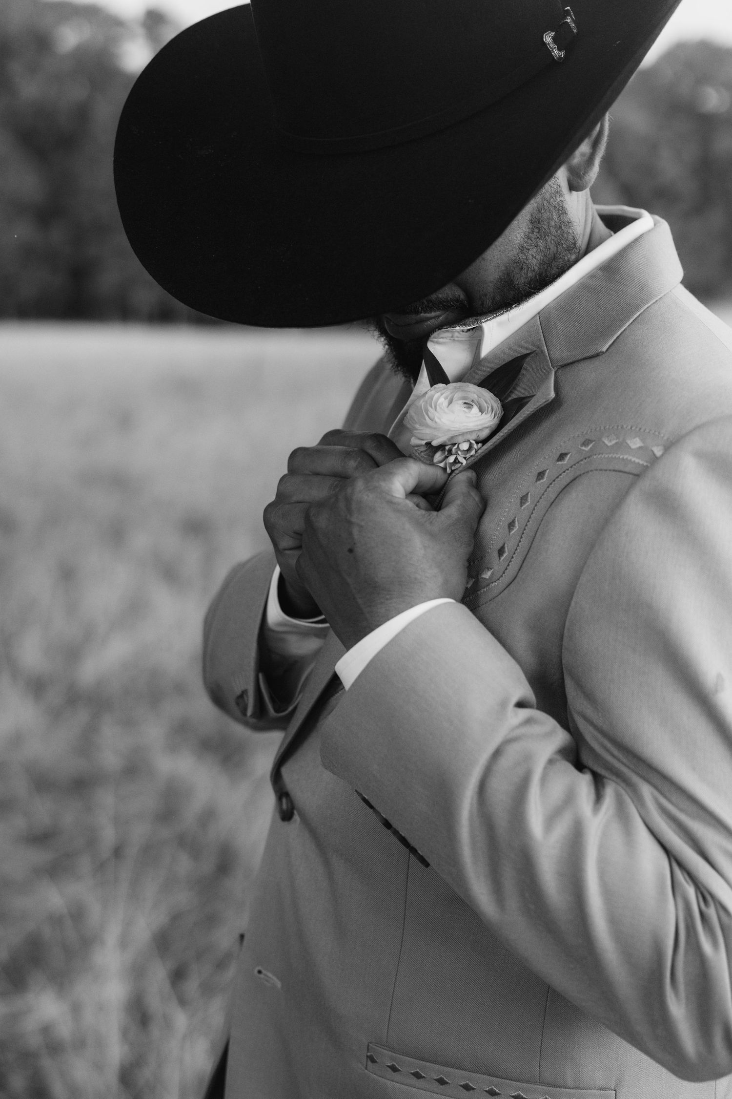 Black and white photo of a groom in cowboy hat adjusting floral boutonniere on suit lapel outdoors.