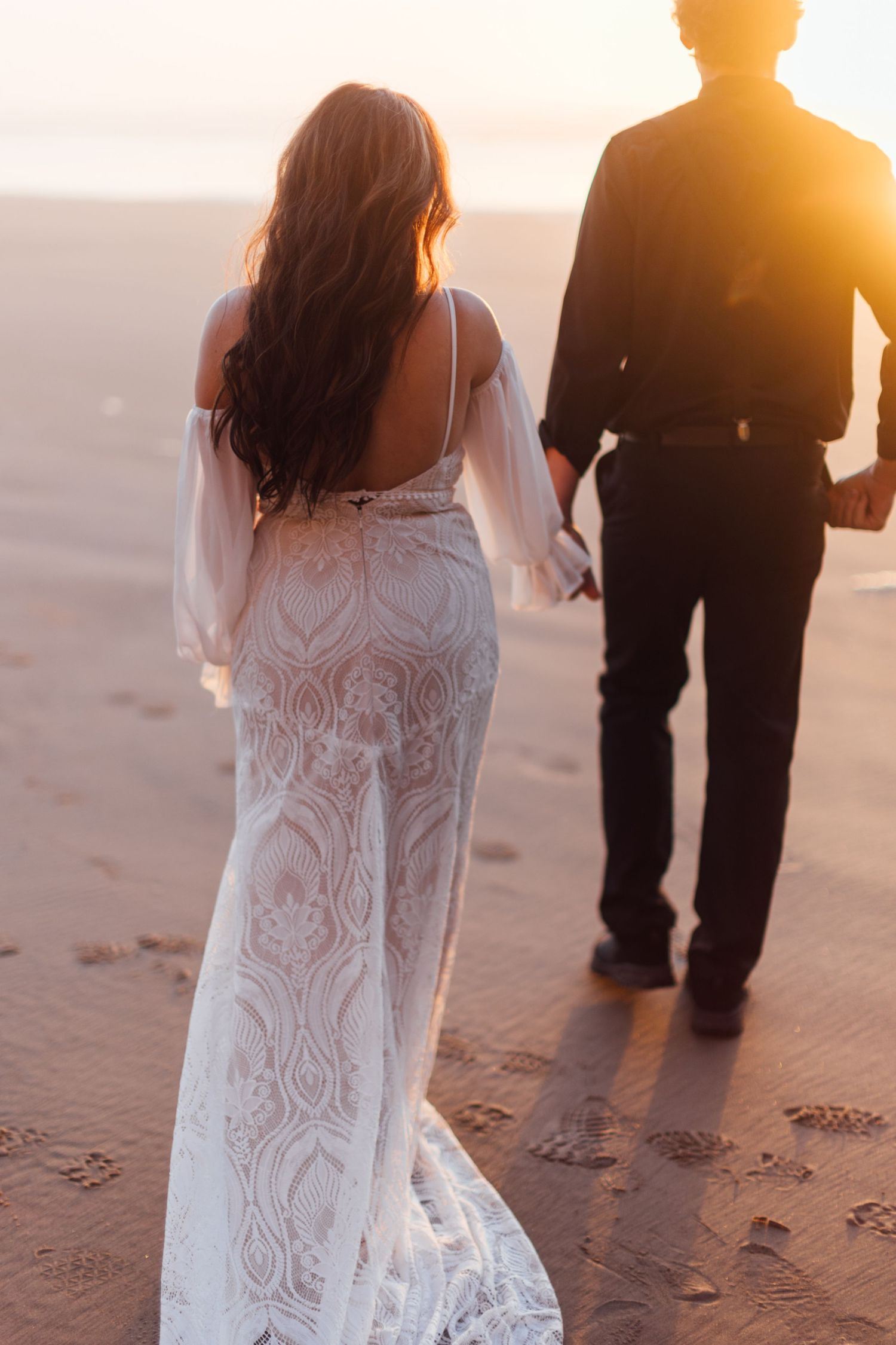 Bride in lace wedding gown holds groom's hand walking on sandy beach at golden sunset.