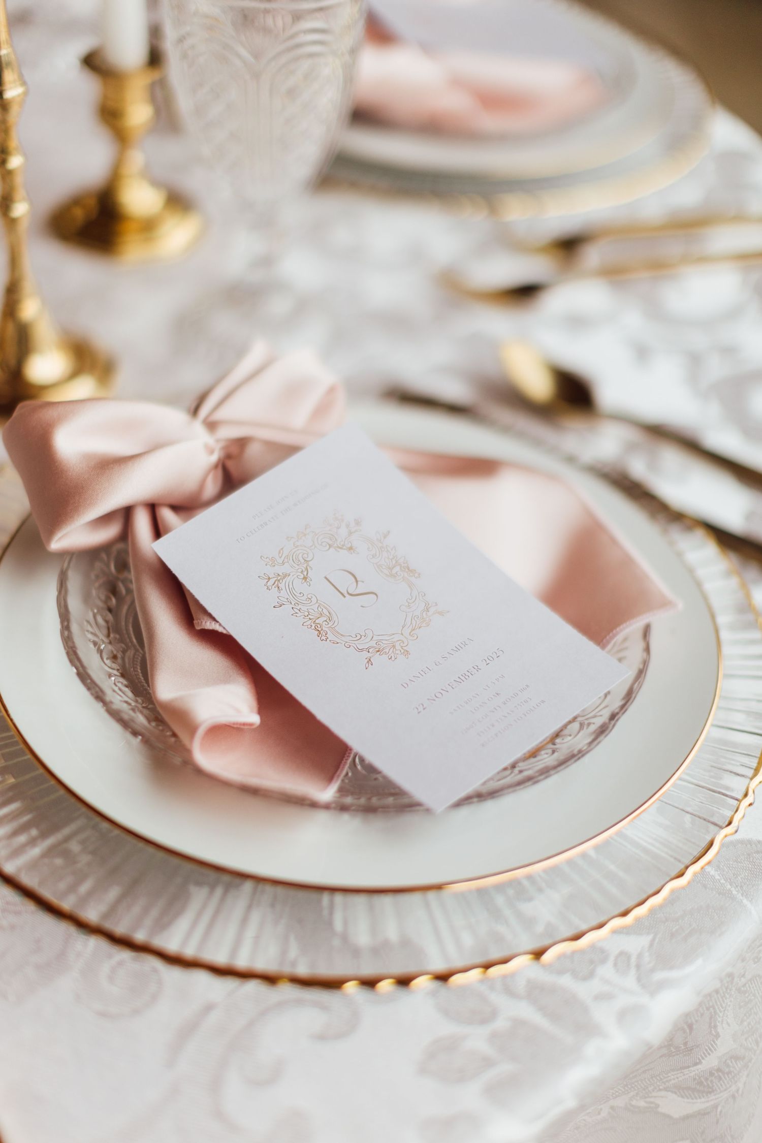 Elegant place setting with pink bow napkin and white invitation card on gold-rimmed plate with lace tablecloth.