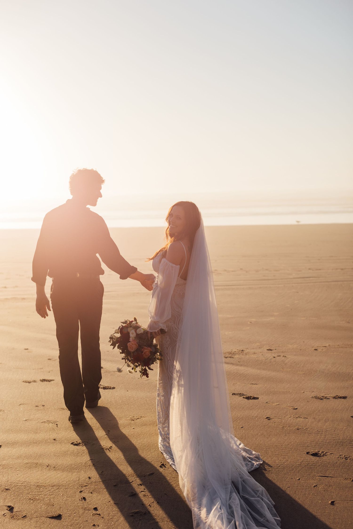 Bride and groom holding hands on a sunlit beach at golden hour, bride wearing a flowing white gown with veil.