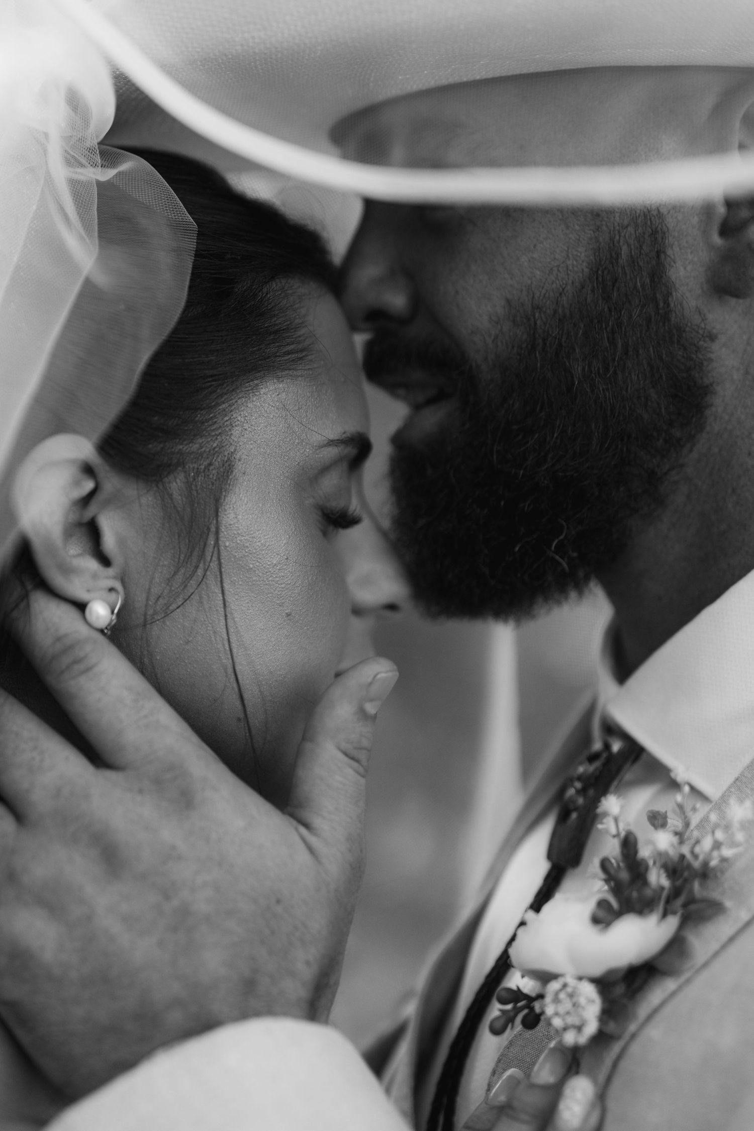 Black and white photo of bride and groom sharing an intimate moment under a veil, groom wearing floral boutonniere.