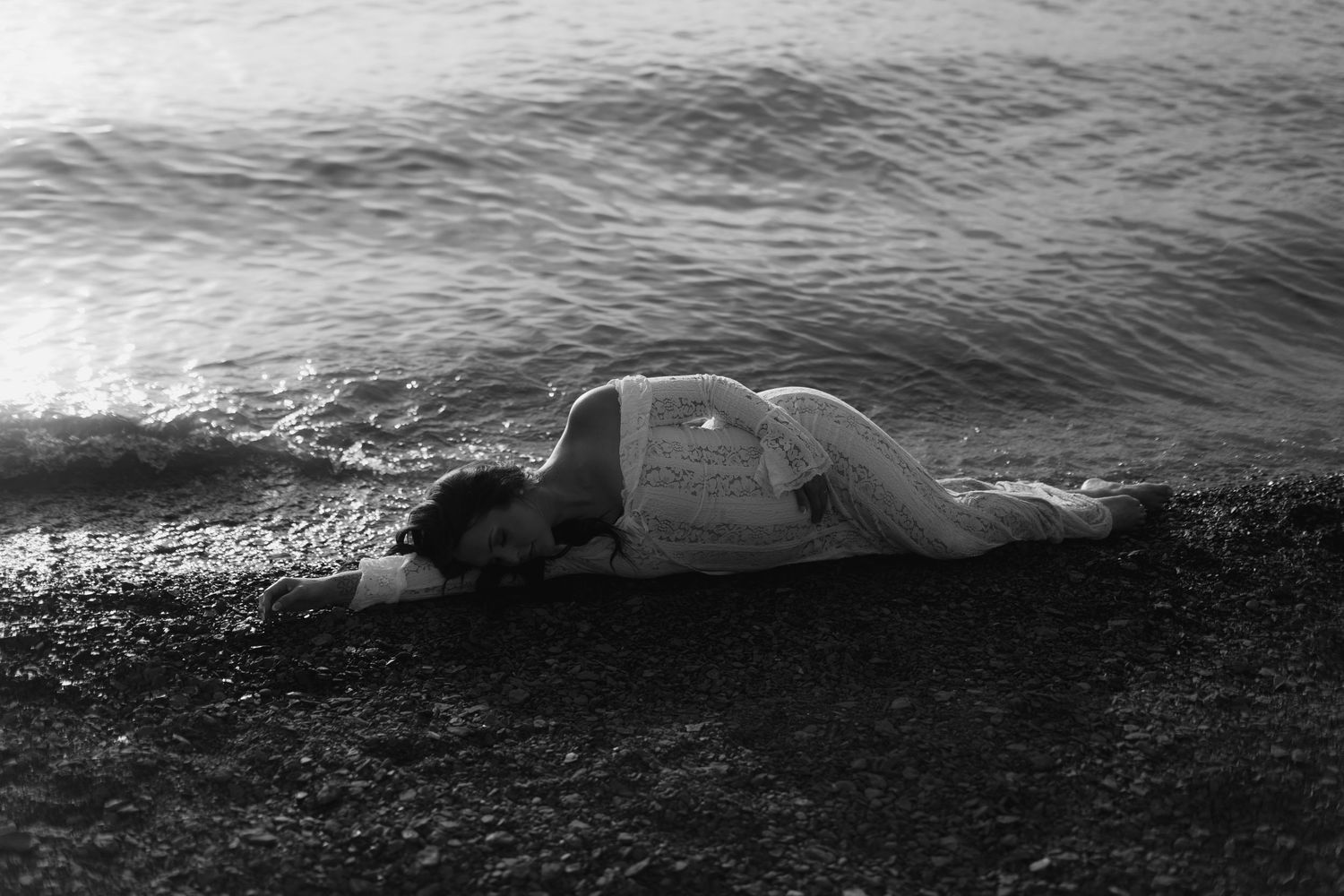 Black and white photo of a woman in white dress lying on rocky shore with gentle ocean waves.