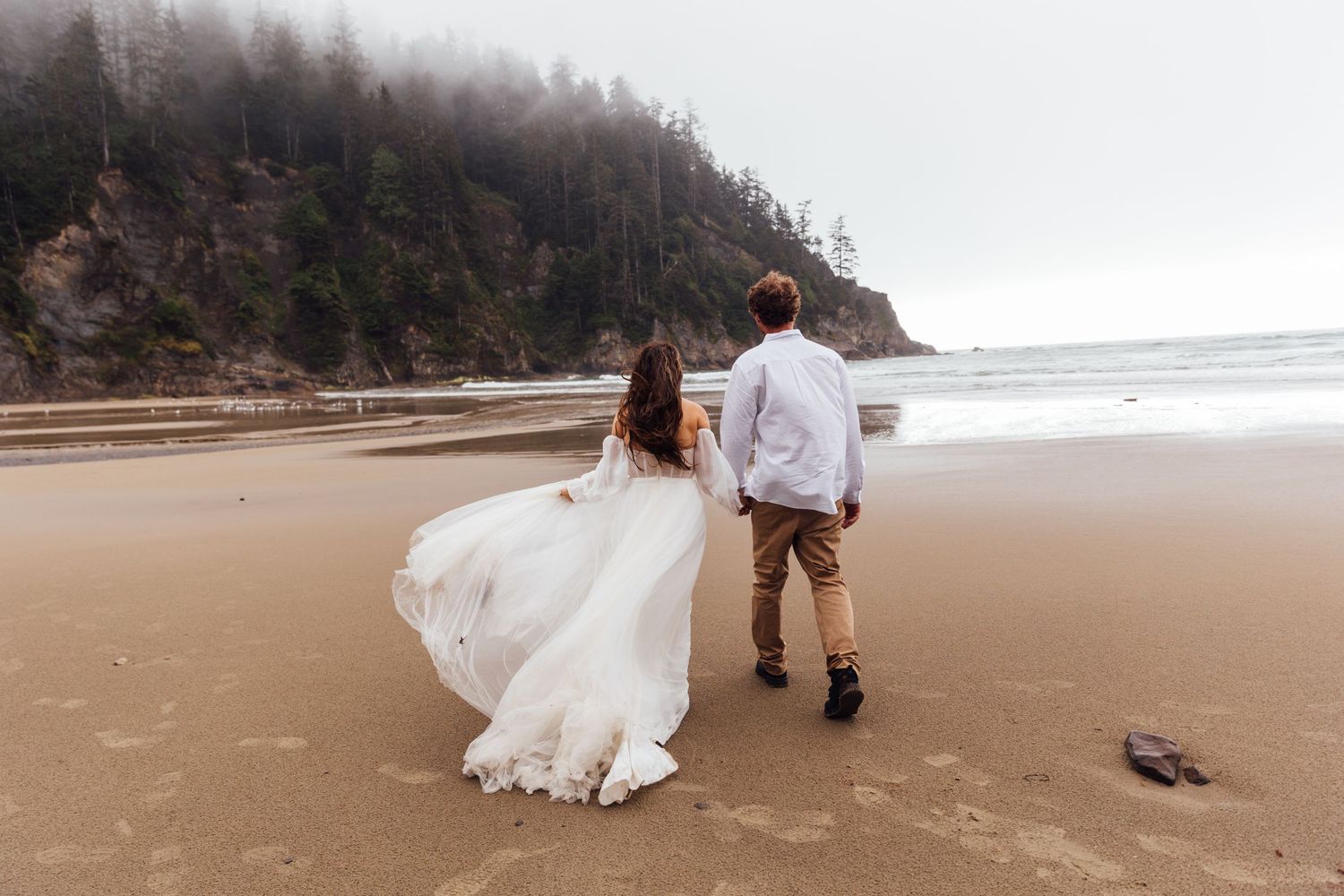 Bride and groom holding hands walking on a misty beach, wedding dress flowing in the wind with forested cliffs behind.