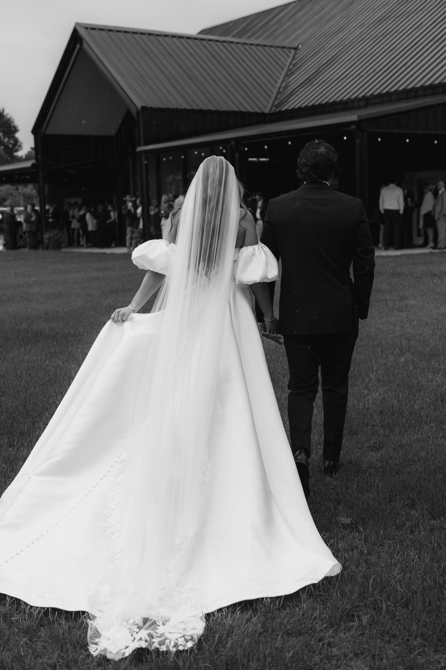 Black and white photo of bride in flowing gown with veil and groom in suit walking toward barn wedding venue.