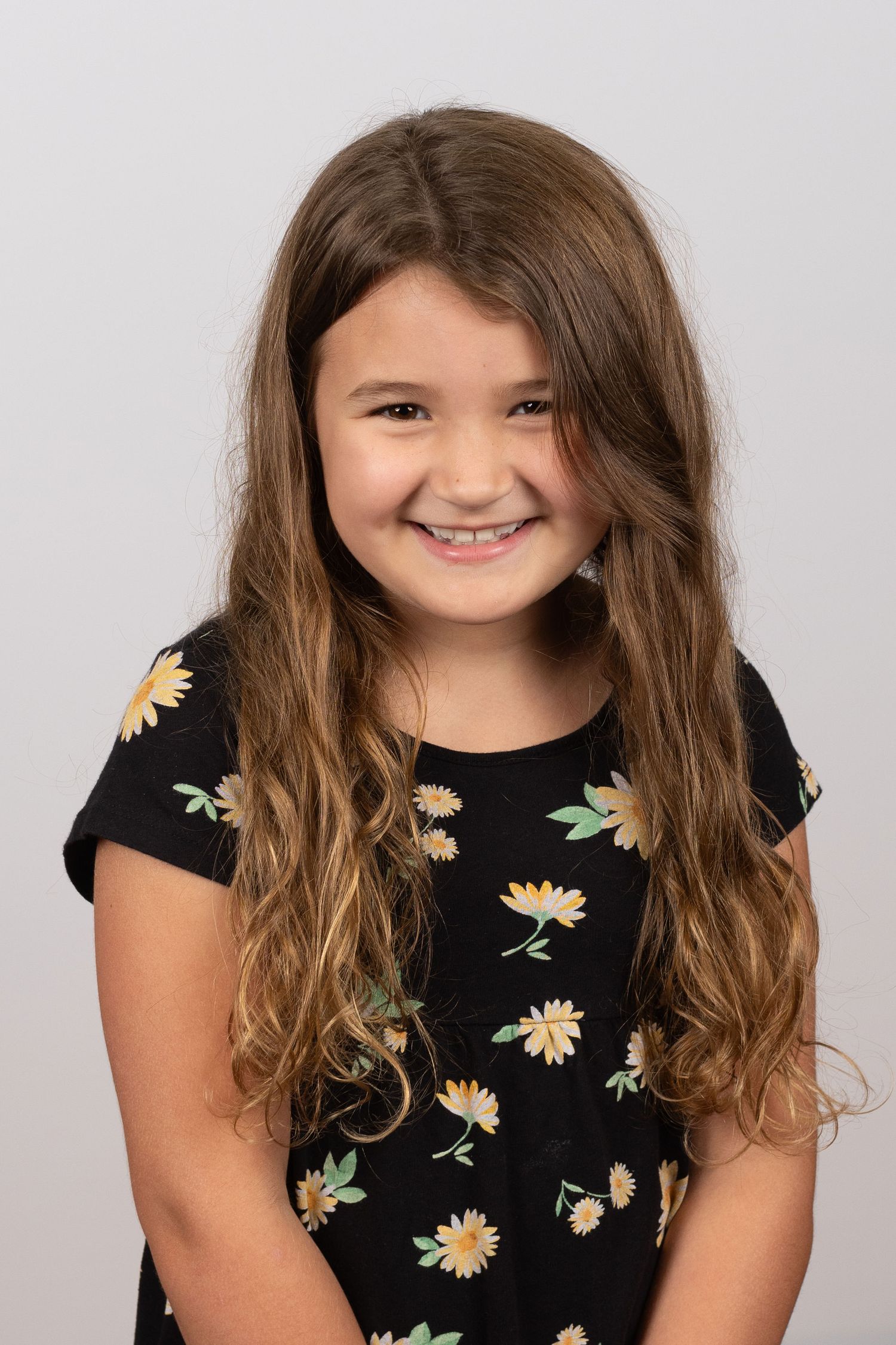 A young person with long wavy brown hair wearing a black floral dress with daisy print smiles at the camera.