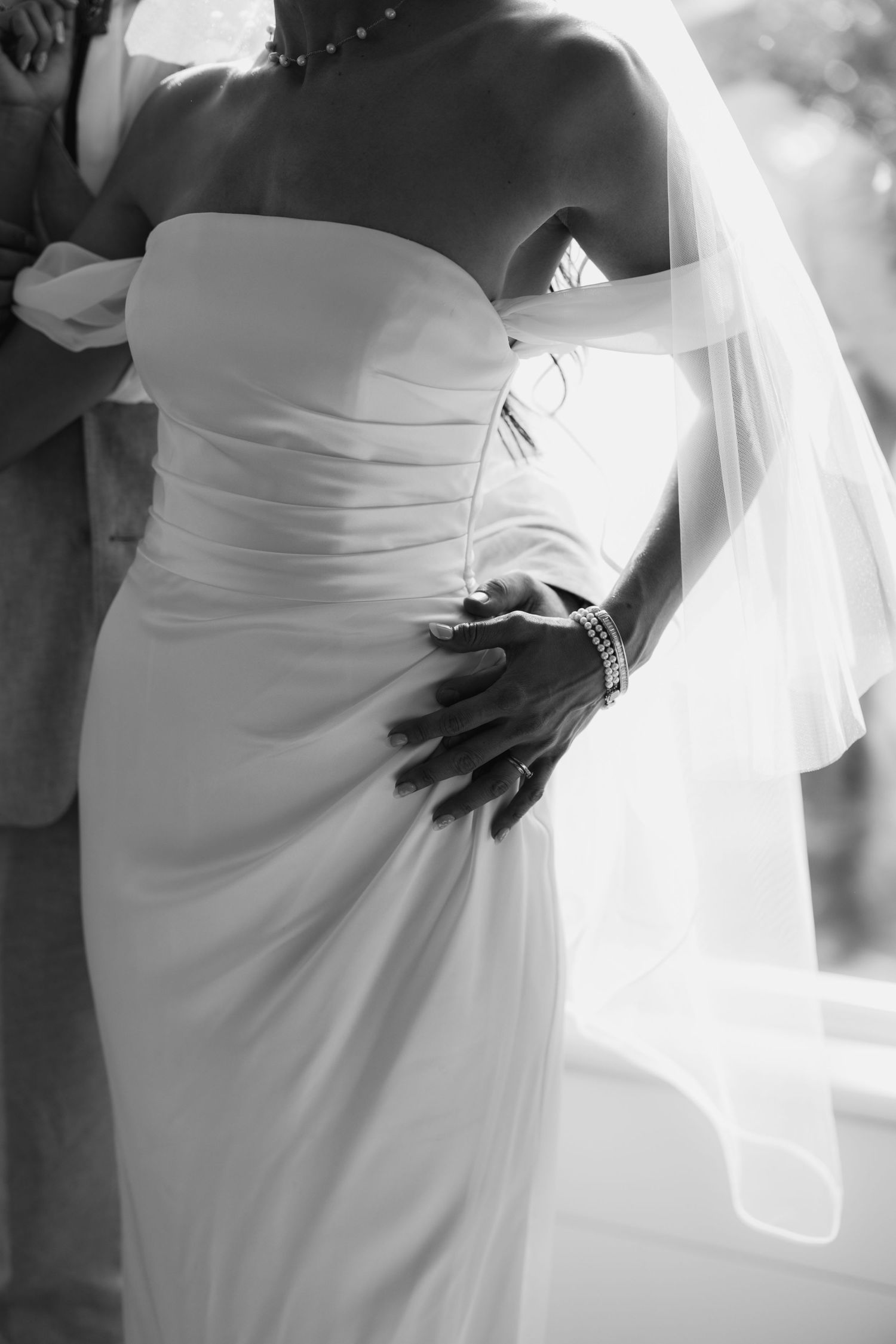 Black and white photo of a bride in a strapless wedding gown with veil, groom's hand on her waist.