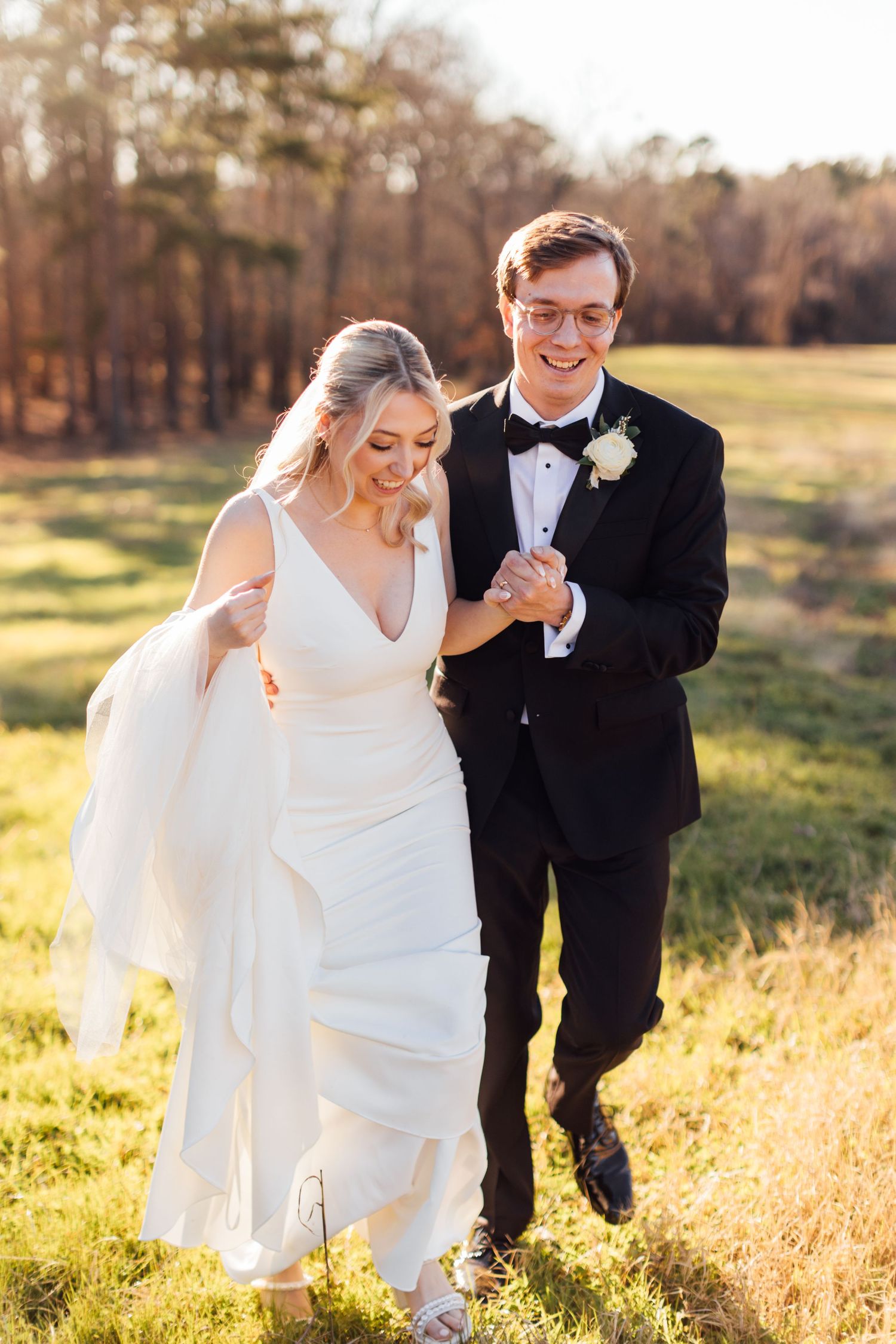 Wedding couple walks hand in hand through sun-drenched field with flowing white dress.
