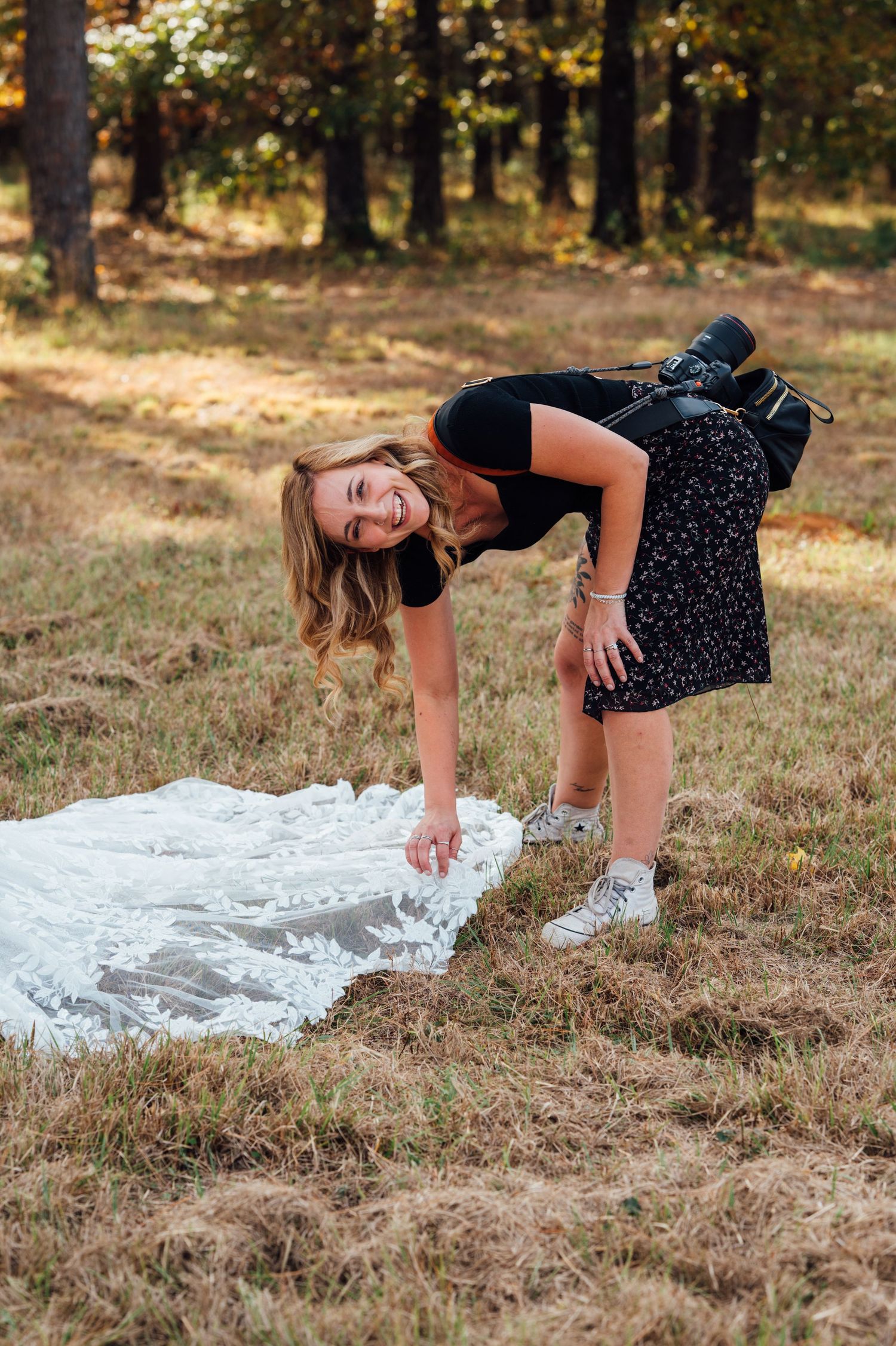 A person in a polka dot dress and sneakers playfully touches a white wedding dress laid out on autumn grass in a forest setting.
