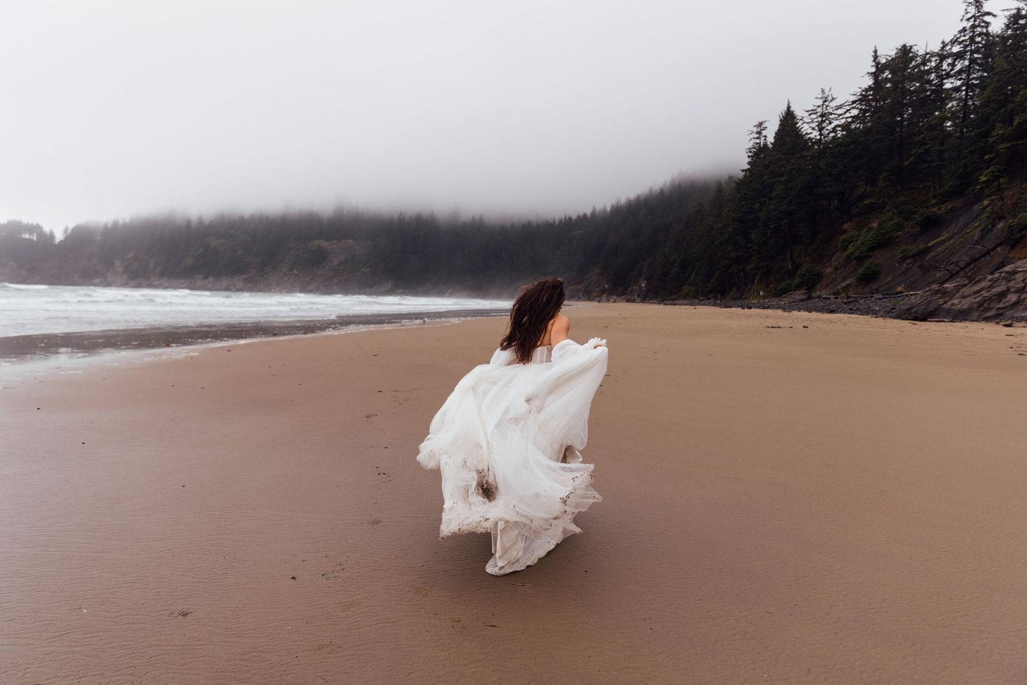 Woman in flowing white dress walking on misty beach with evergreen trees and fog in background.
