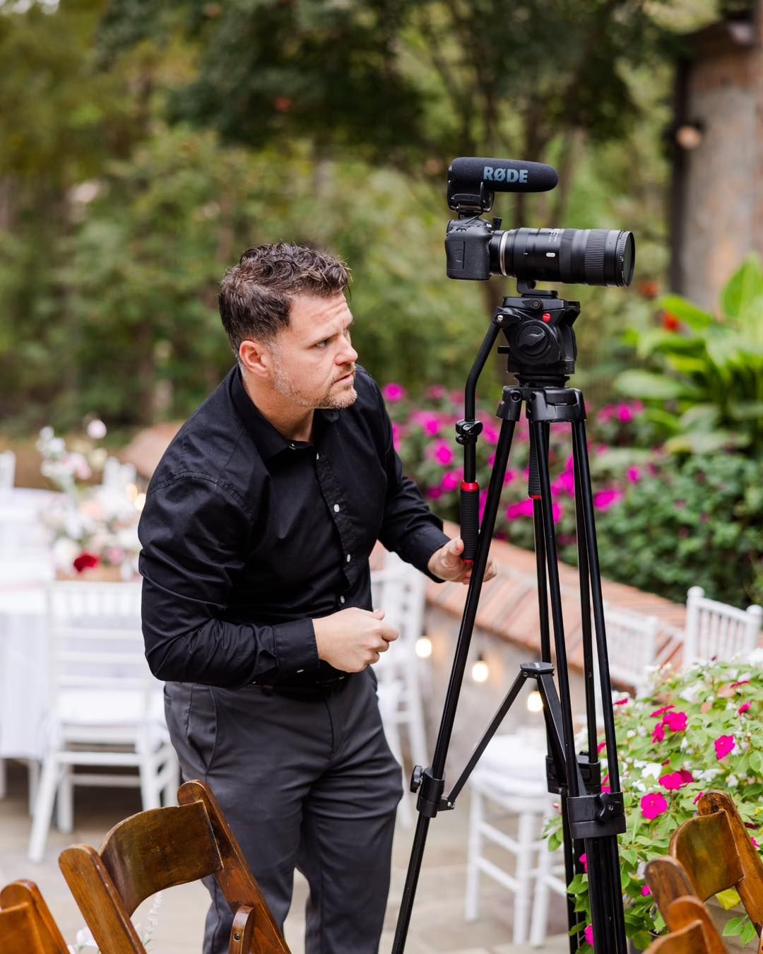 Professional videographer with camera on tripod filming at an outdoor event with white chairs and pink flowers in background.
