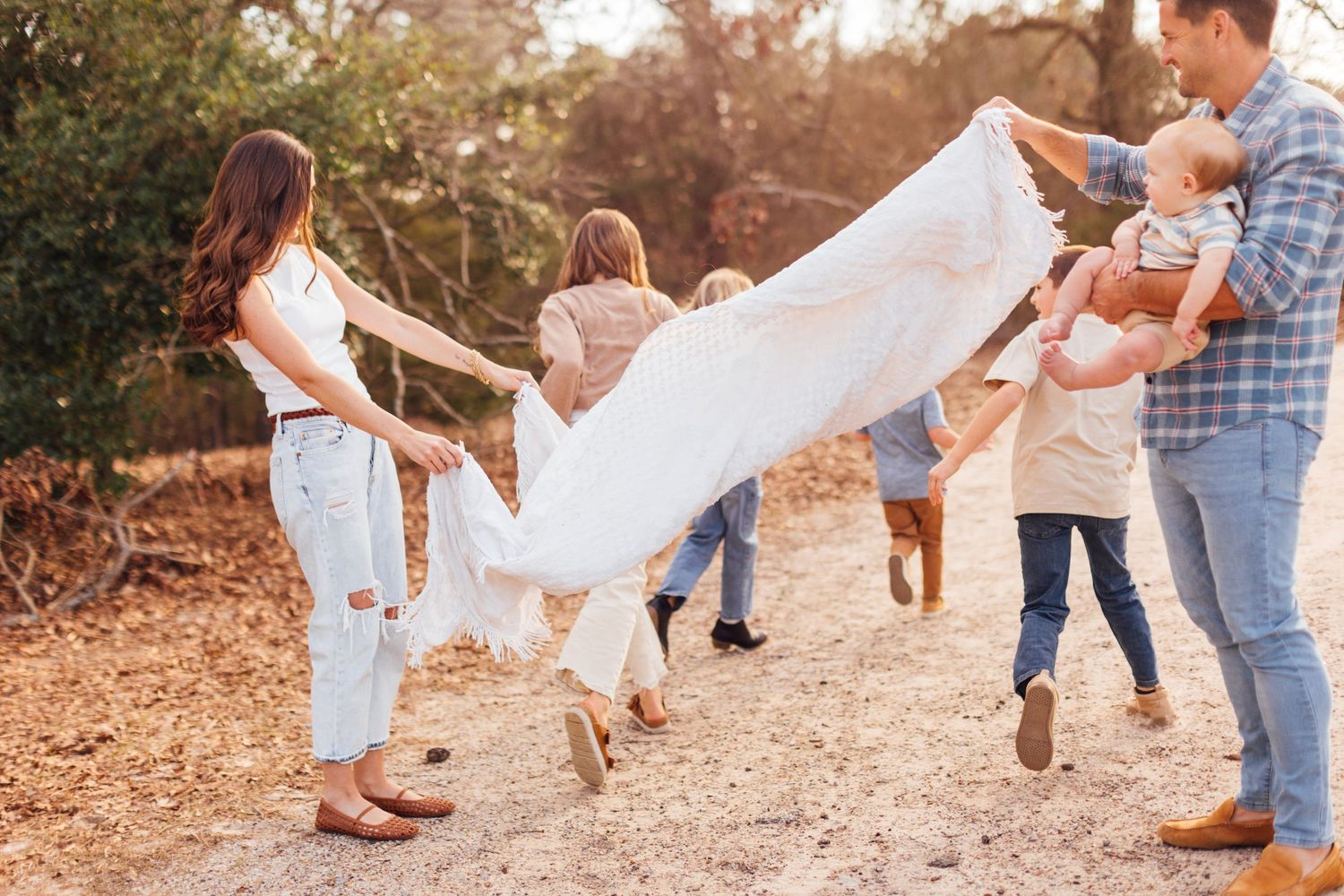 Family playfully shaking a white blanket outdoors on a sunny dirt path surrounded by autumn trees.
