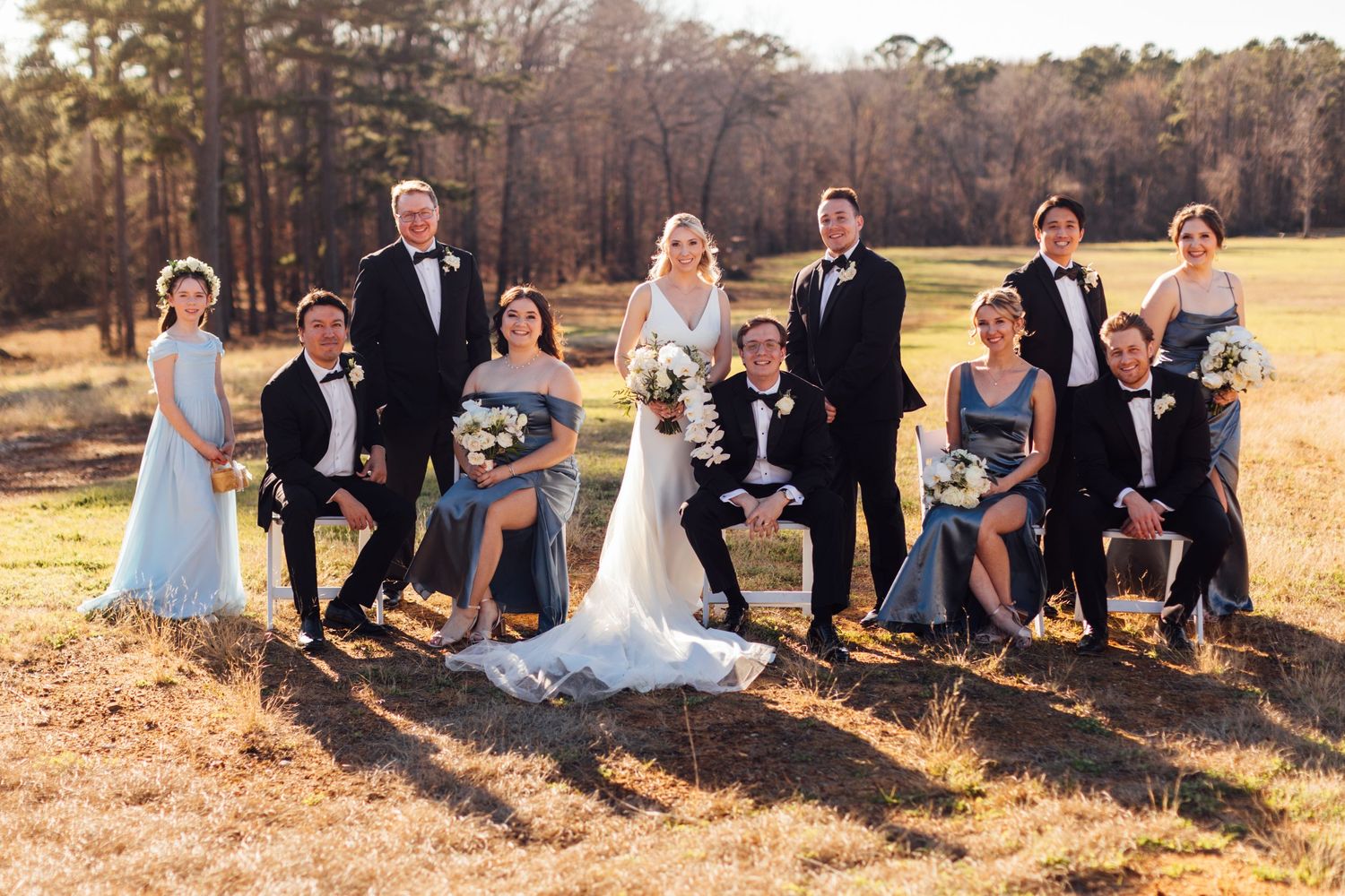 Wedding party relaxes on the grass in front of autumn trees during sunset photography session.