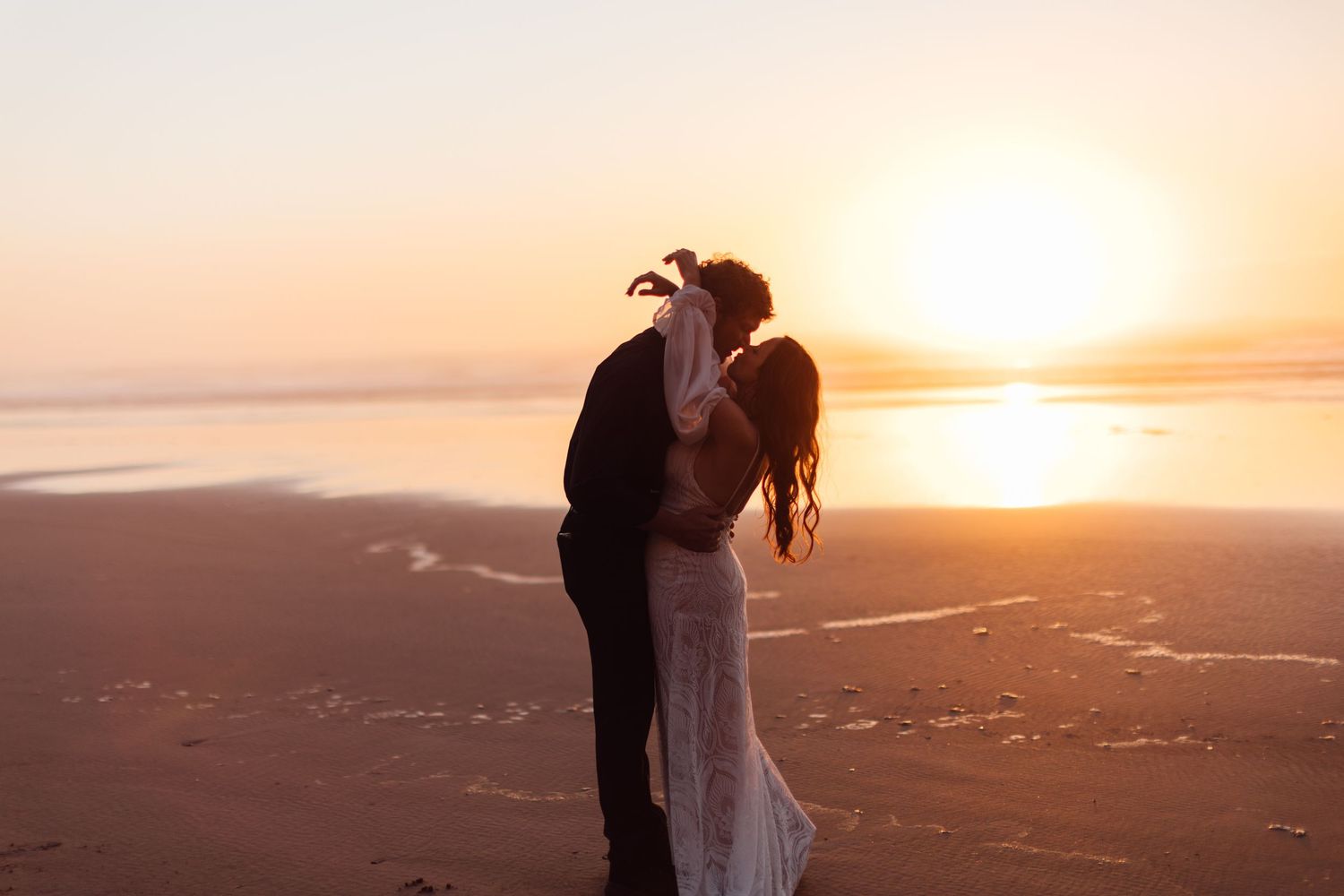 Couple sharing a romantic kiss on a sandy beach at golden sunset, wedding attire, ocean backdrop.
