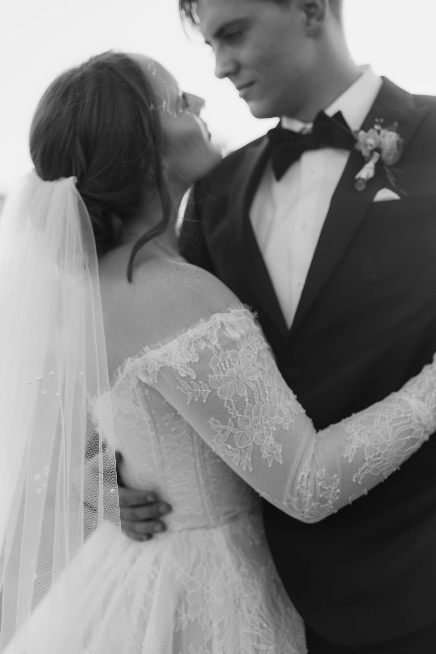 Black and white photo of bride in lace gown and veil embracing groom in tuxedo at wedding.