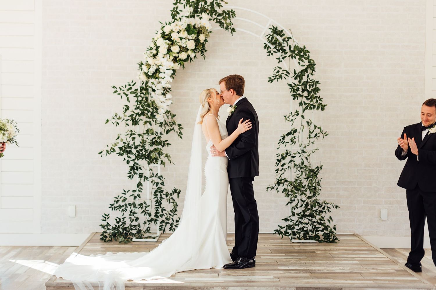 Wedding couple shares romantic moment against white wall adorned with floral greenery.