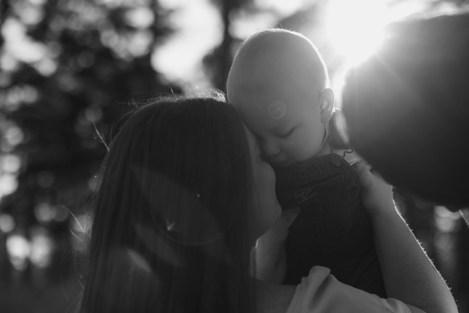 Black and white photo of a mother tenderly holding her baby up close, with sunlight glowing softly behind them.