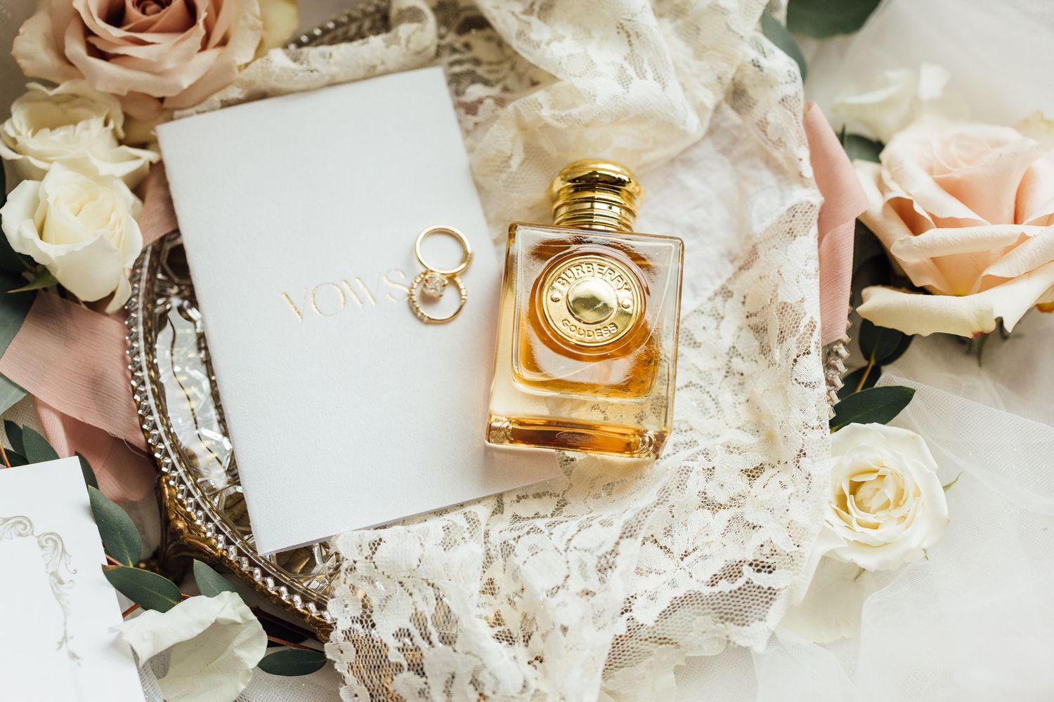 Wedding rings and perfume bottle arranged with blush roses and lace on white background.