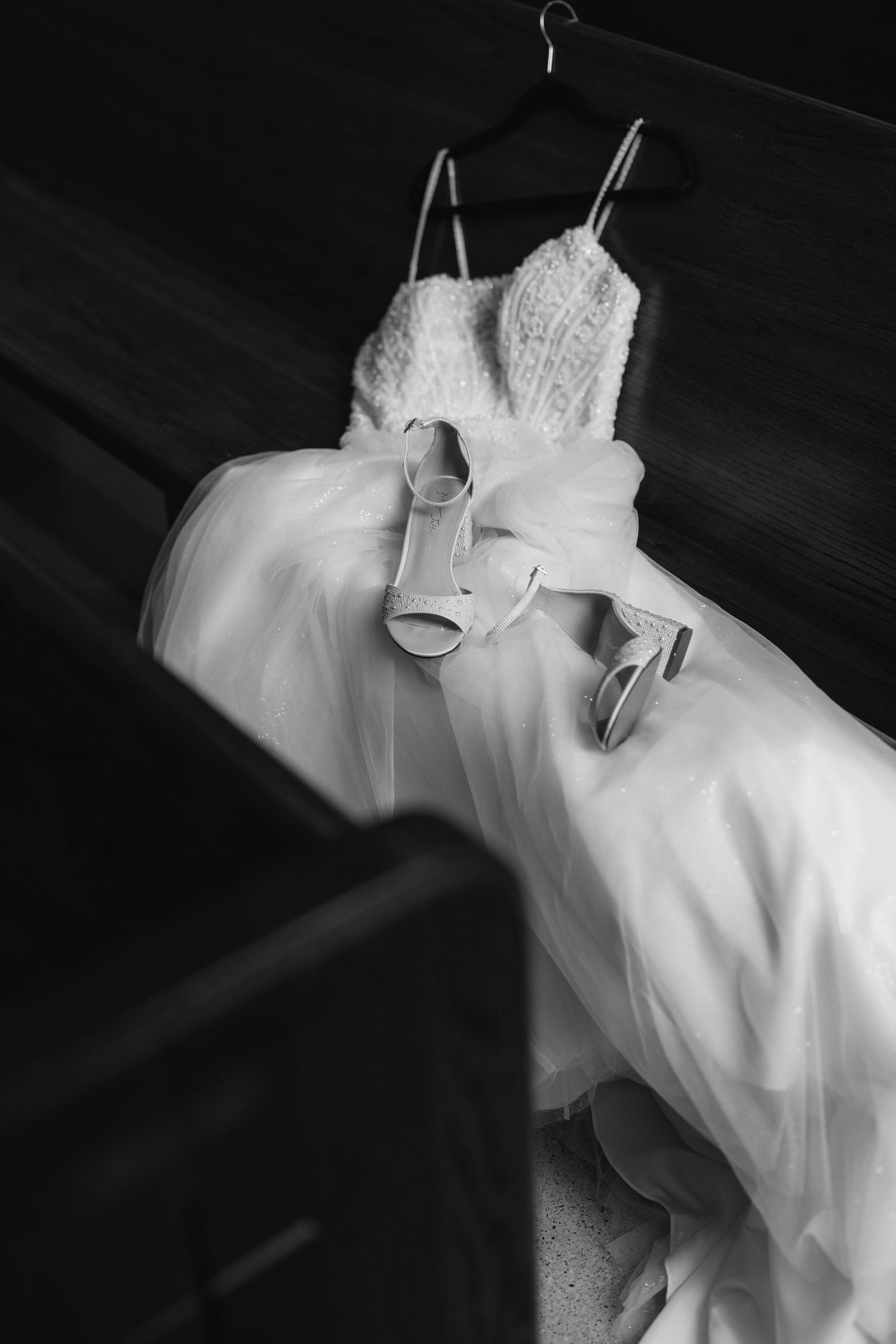 Black and white photo of a wedding dress hanging with bridal heels, elegant lace bodice and flowing skirt on display.