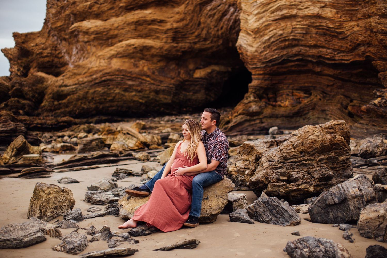 Couple sitting together on rocky beach with dramatic sandstone cave formation backdrop during engagement photo session.