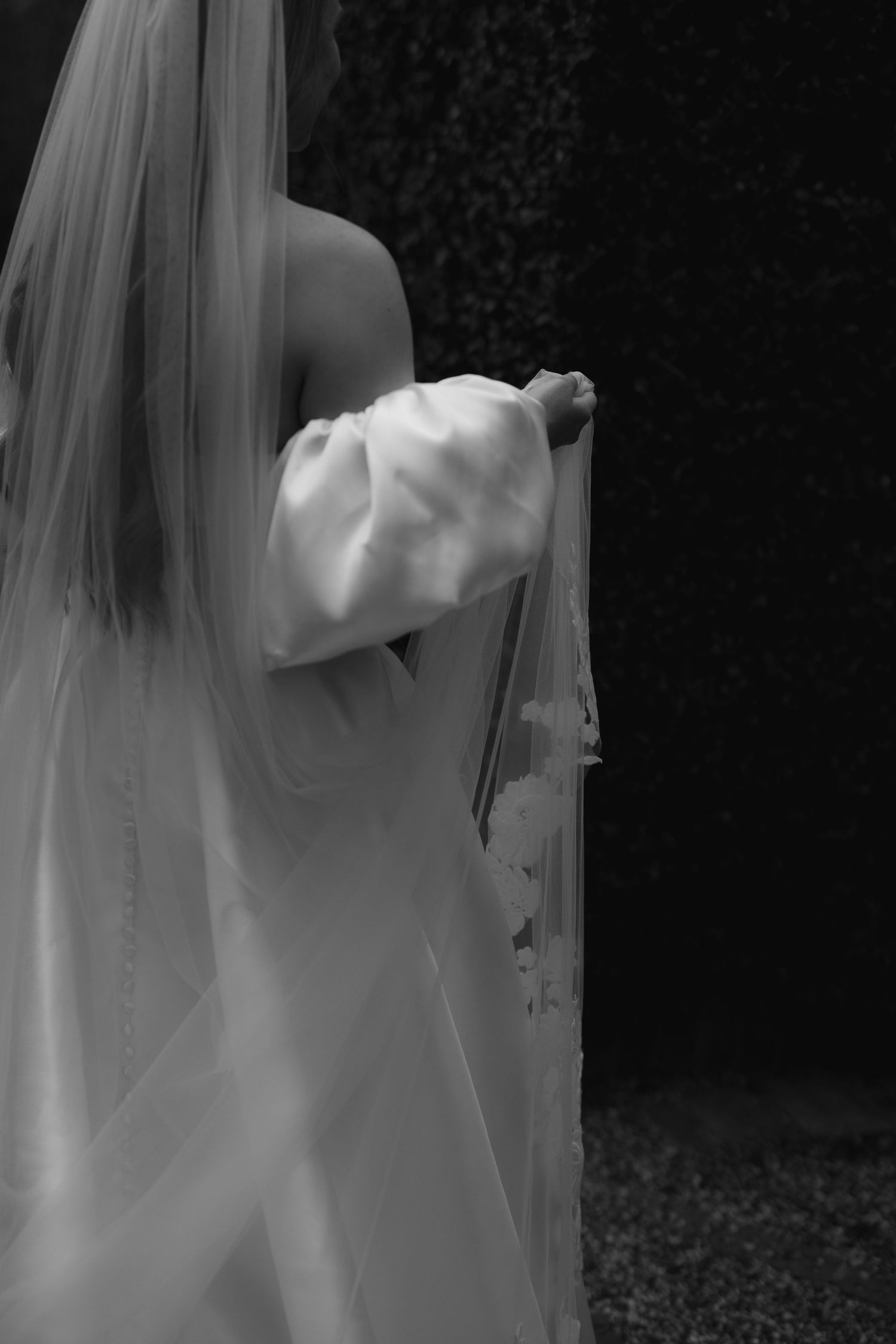 Black and white photo of a bride in a white gown with puff sleeves and long veil, viewed from behind outdoors.
