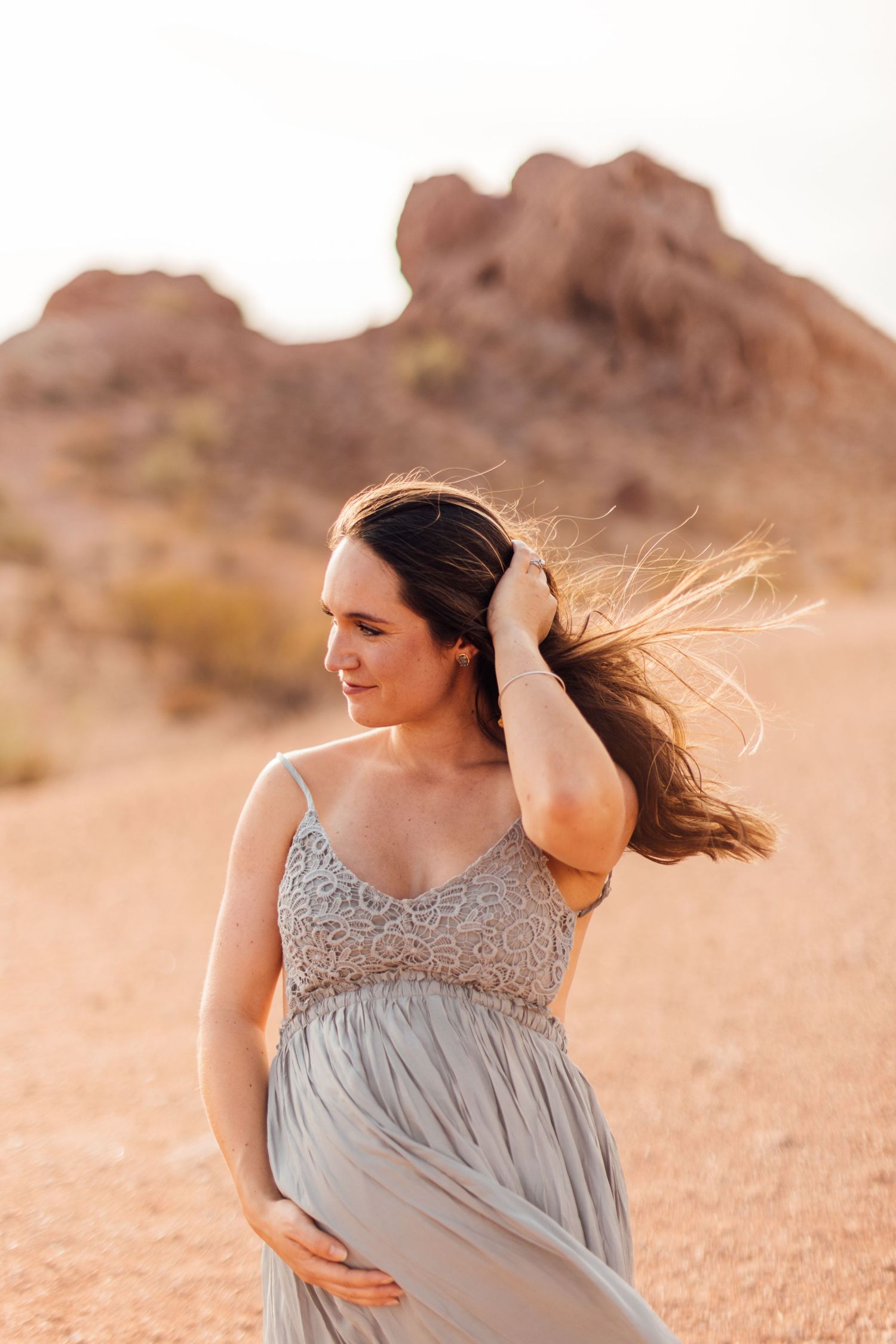 Pregnant woman in gray dress holds hair in desert wind, rocky formations behind her in warm sunset light.