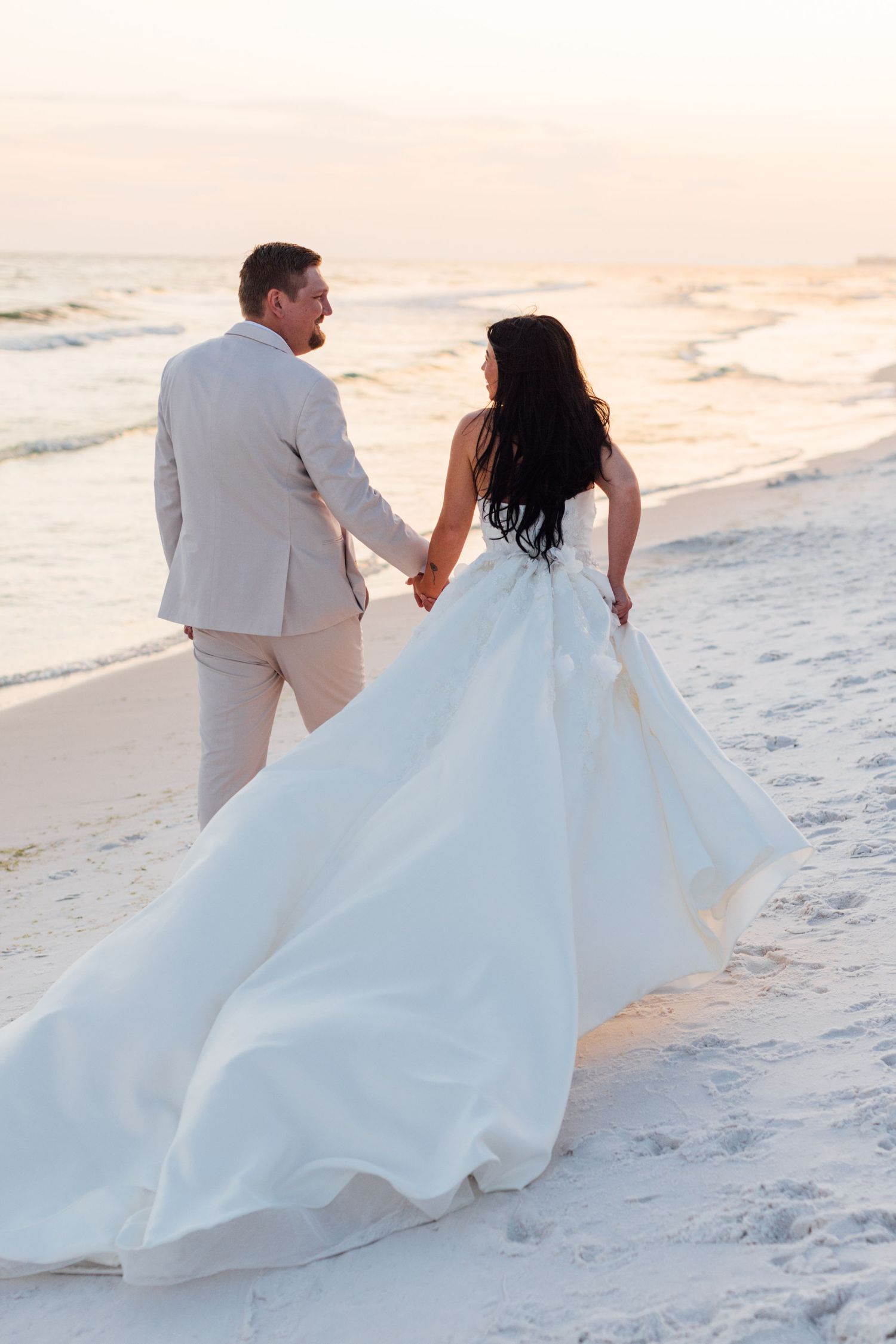 Wedding photography and groom holding hands walking on a white sand beach at sunset, wedding dress train flowing behind them.