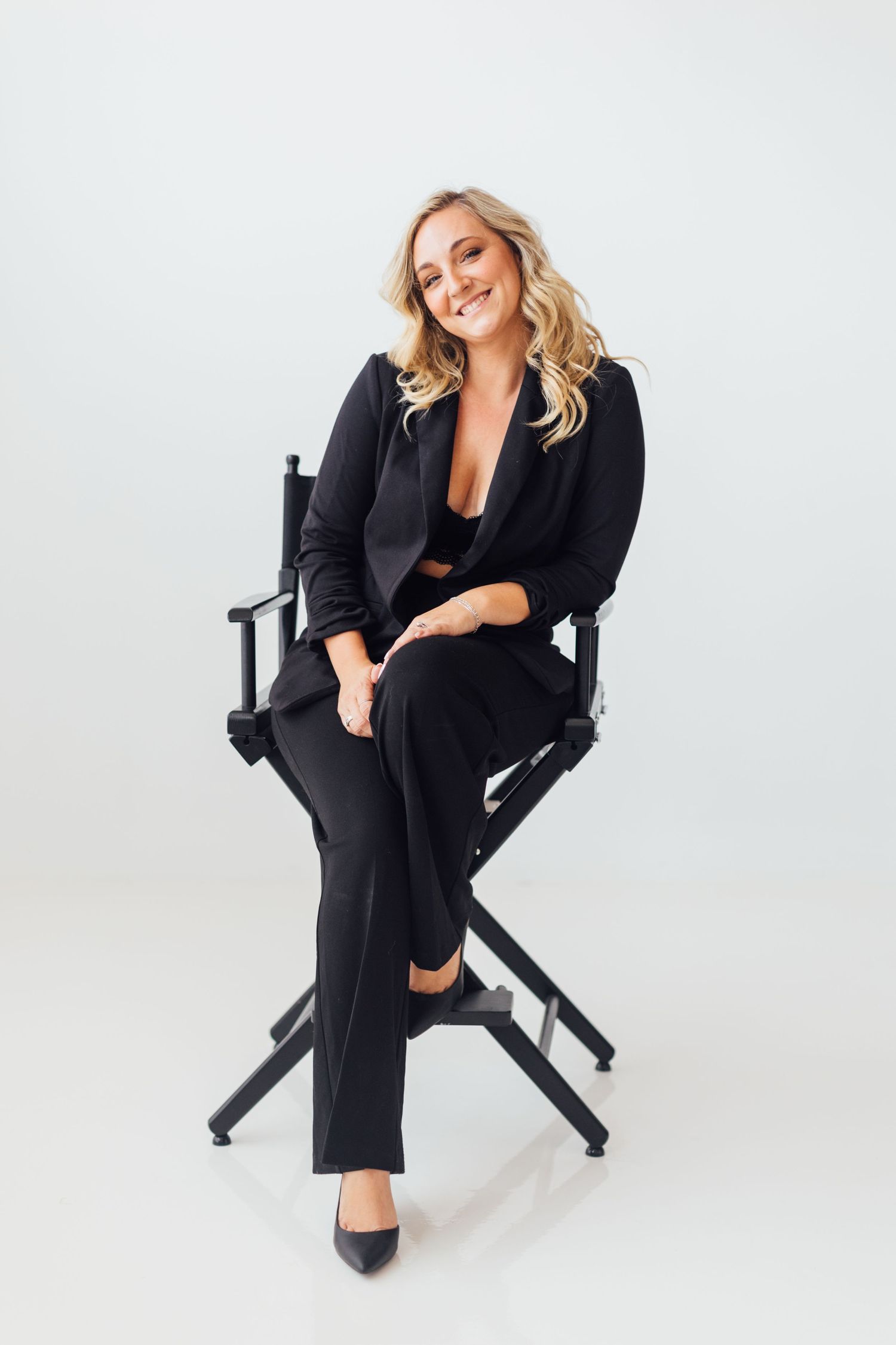 Professional wearing all black business attire poses confidently on director's chair against white backdrop in studio setting.