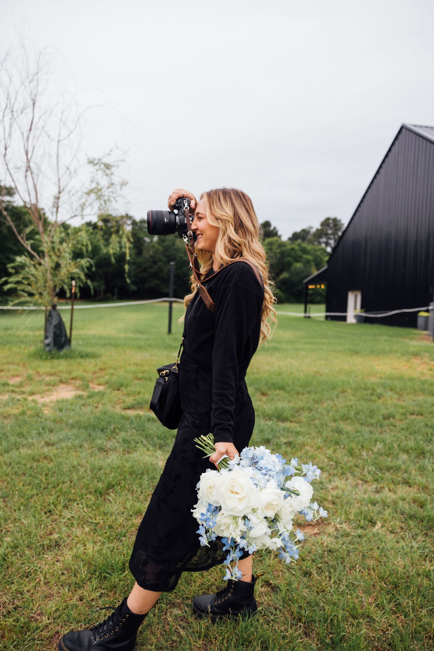 Wedding Photographer of East Texas londe woman in black dress holding blue and white flowers while photographing outdoors near a black barn.