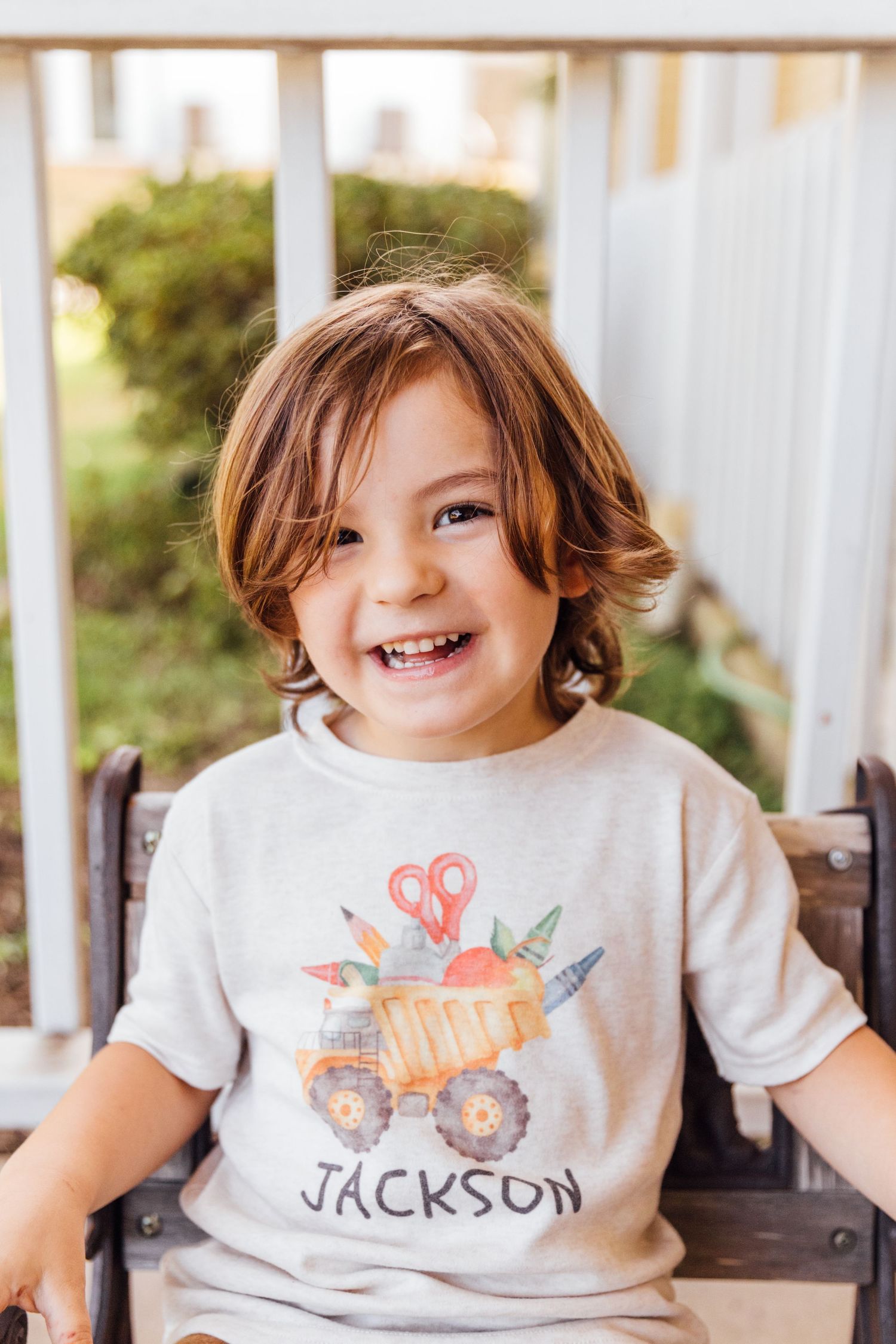 Child sitting on porch wearing light gray t-shirt with colorful truck design and name Jackson.
